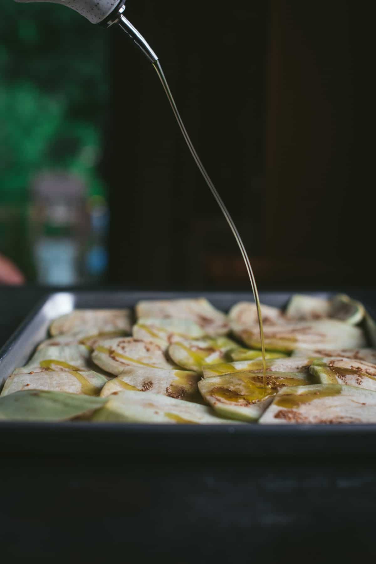 A hand pours olive oil from a height to land on top of the resting eggplant. The eggplant are resting in a silver baking tray and are overlapping slightly.