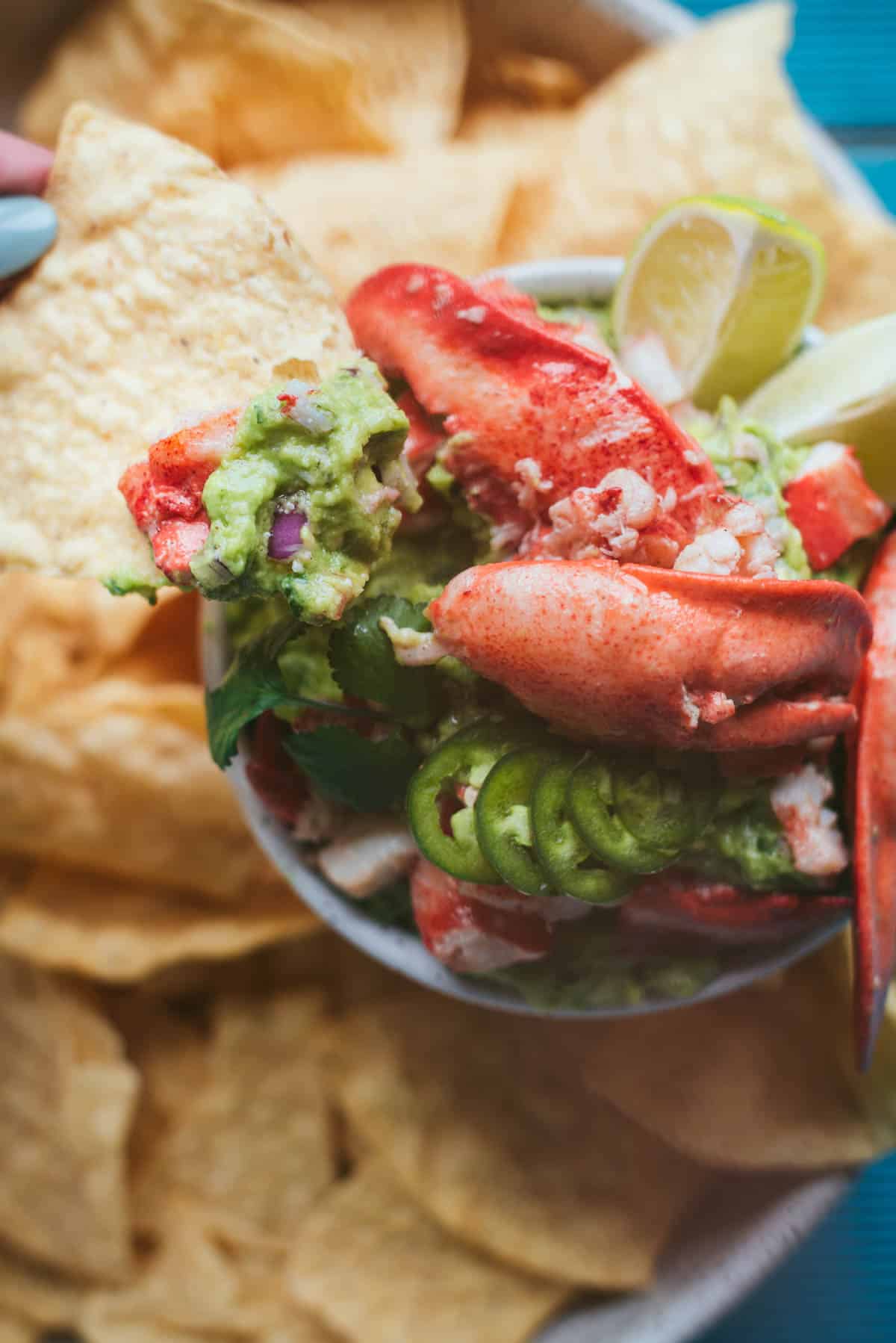 Close up birds eye view of lobster guacamole. You can see all the small details like the chopped up red onion throughout the guacamole. The lime wedges are there and the lobster is bright red and in the center of the bowl and main focus of the picture. You can see the chips for dipping around the edges of the photo, slightly out of focus.