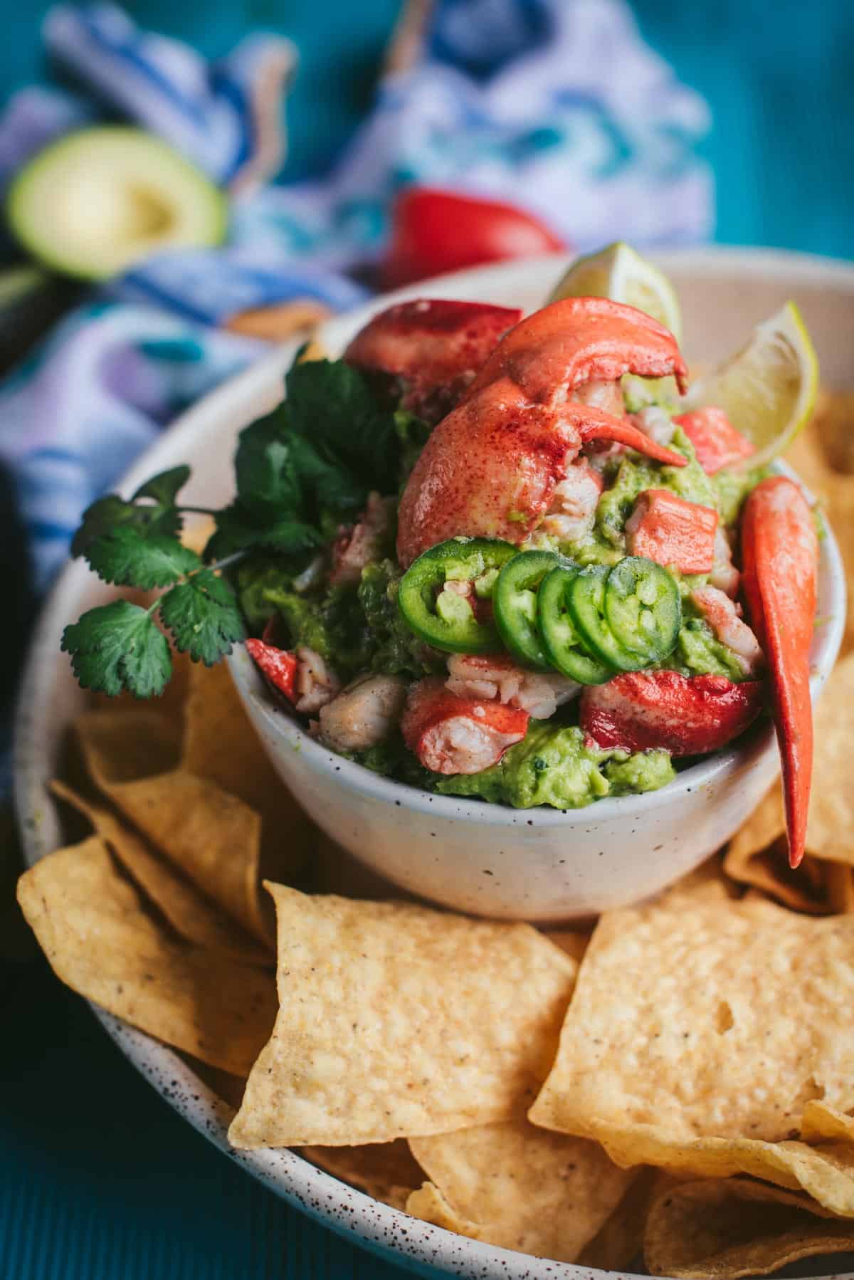Close up view of lobster guacamole. The red lobster tails are sitting on top of the guac and there is jalapenos and cilantro on top of that. You can see the chips for dipping on the plate at the bottom slightly out of focus.