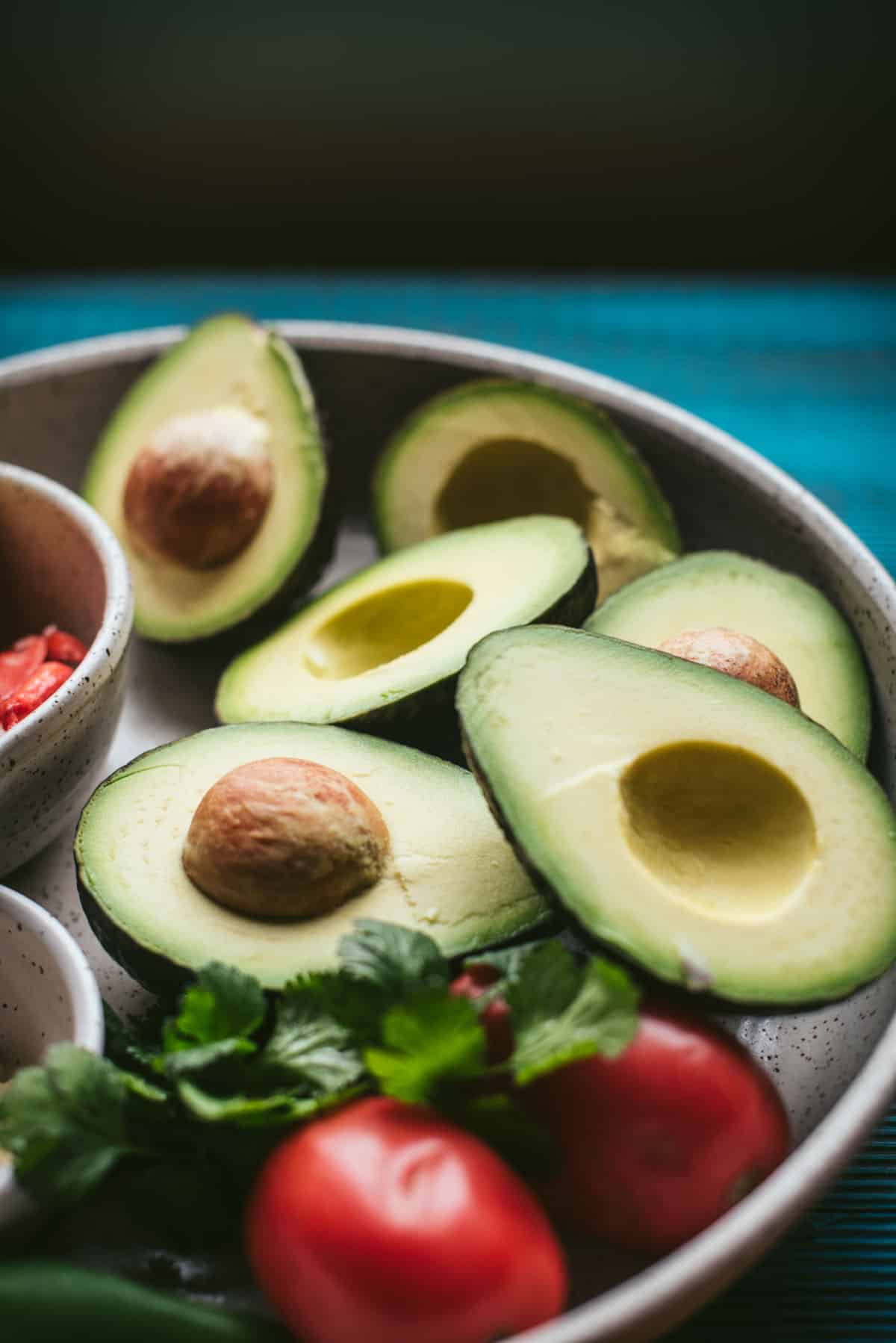 Close up of ingredients, 3 avocados sliced in half, with the seeds still intact inside the avocado half. There are 2 tomatoes and a small hand full of cilantro.