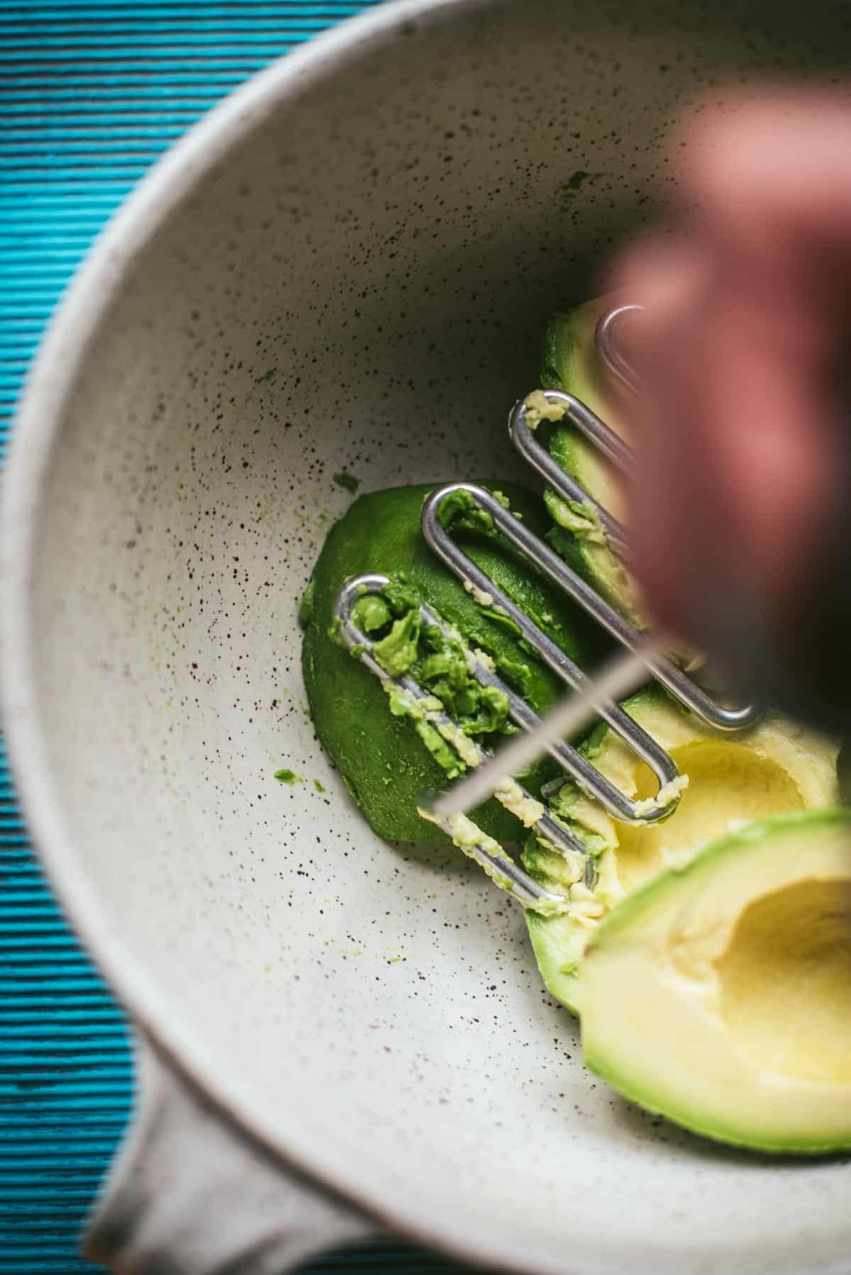 Process picture, mashing the avocado in a white terrazzo mixing bowl with a metal masher.