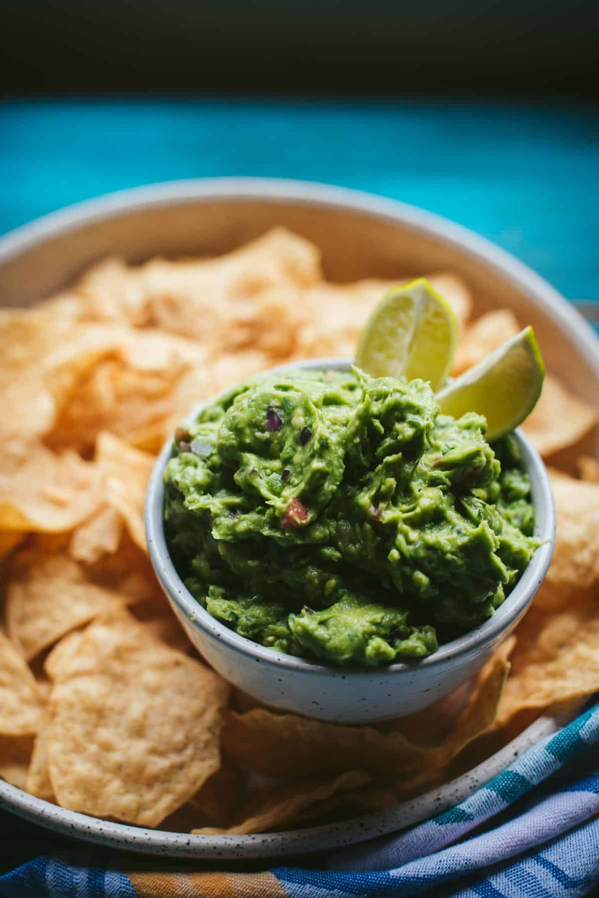 The guacamole is in the serving bowl with lime wedges added. The bowl is sitting on the white terrazzo plate with chips.