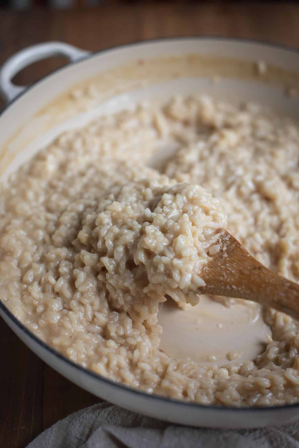 A close up shot of the finished risotto. The rice looks shiny in the white pan. The wooden spoon has scoped up a big piece of risotto and you can see the white of the pan beneath it.