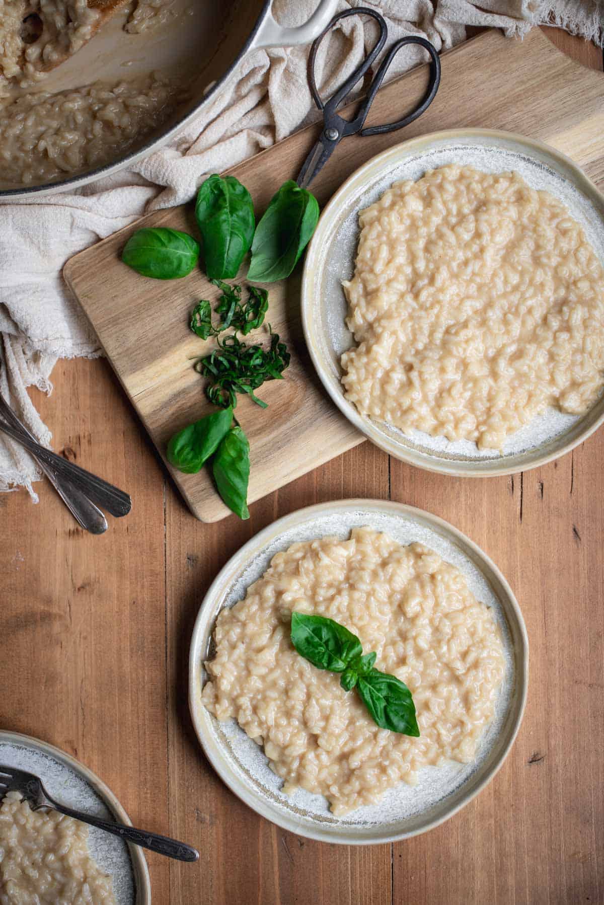 2 grey plates with Parmesan risotto and fresh basil leaves, on a wooden chopping table. There are full and chopped fresh basil leaves on a wooden chopping board. 
Part of the left over risotto in the pan can be seen in the corner.