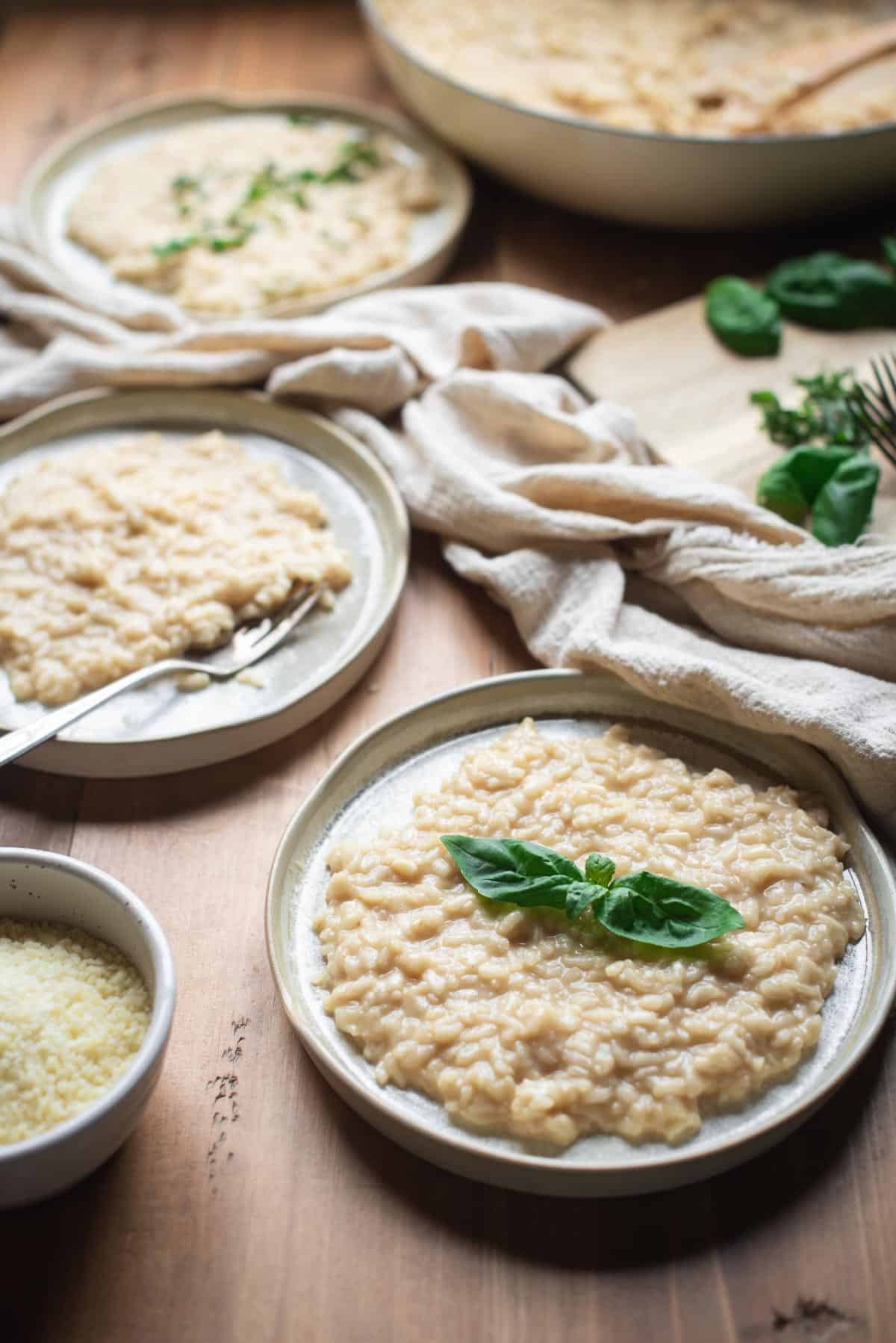 3 neutral colored plates containing parmesan risotto on a wooden table in a line. The empty pan that was used to cook the risotto is sitting at the back out of focus. There is a wooden chopping board with some fresh basil on top, and a bowl of parmesan cheese. One of the dishes has also been topped with fresh basil leaves.