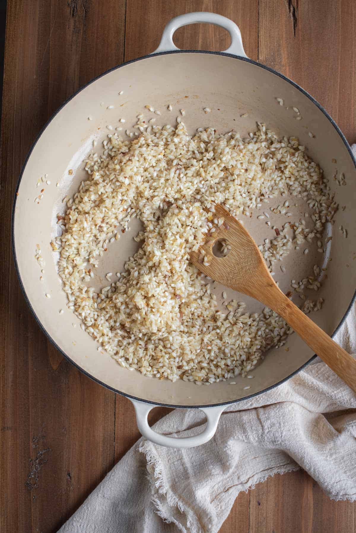 There is a large white skillet with part cooked rice and a wooden spoon on a wooden surface.