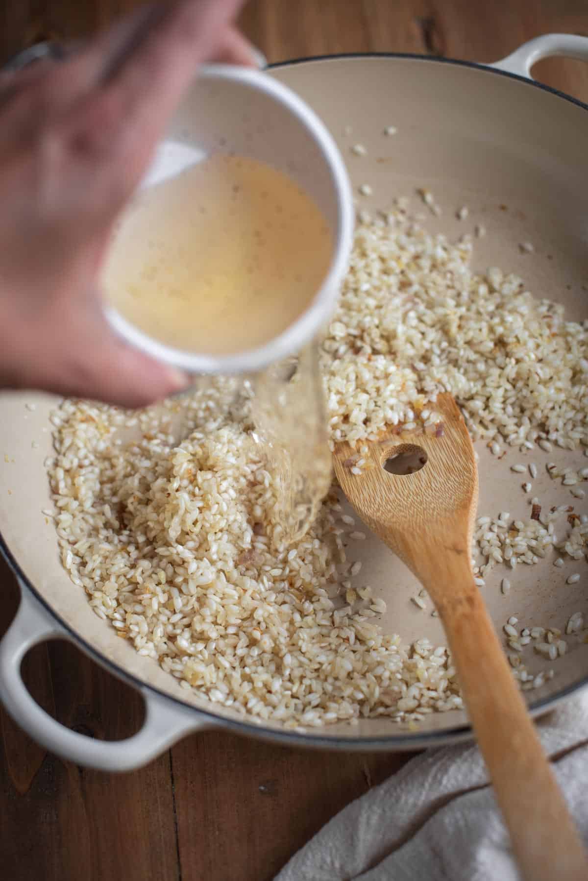 A hand pours white wine from a white cup into the large white skillet containing the cooking rice and shallots. A wooden spoon is resting inside of the pan.