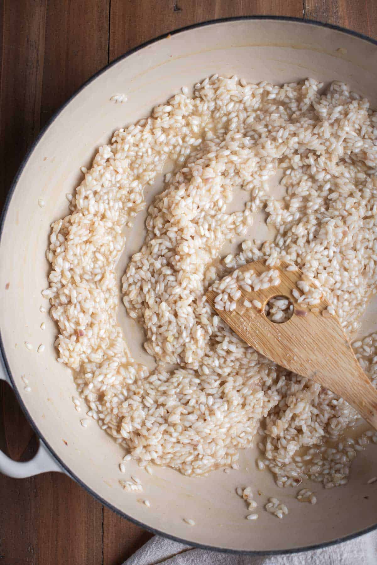 The rice inside the white skillet has increased in size and looks more cooked. The wooden spoon is stirring the rice.