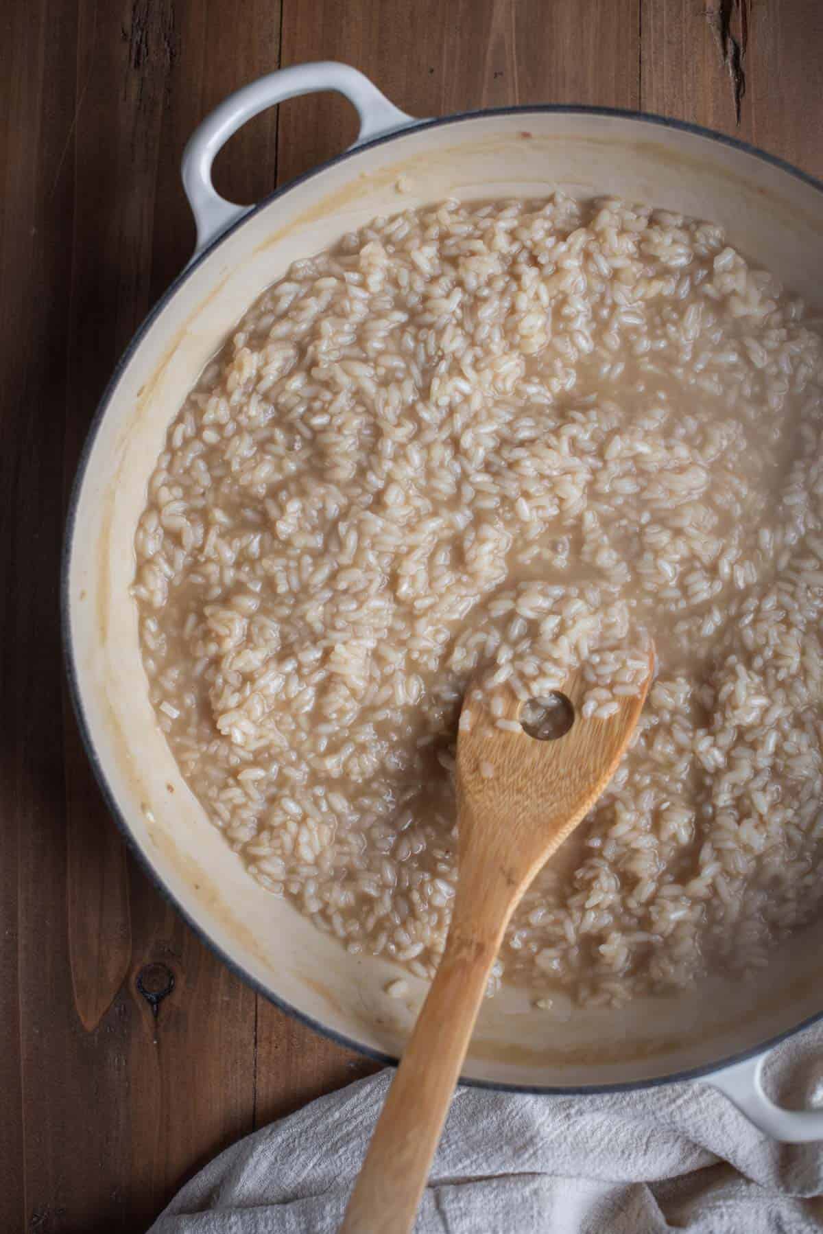A white skillet containing cooking risotto. The mixture looks quite liquidy at this stage. A wooden spoon is lifting some of the mixture out of the pan.