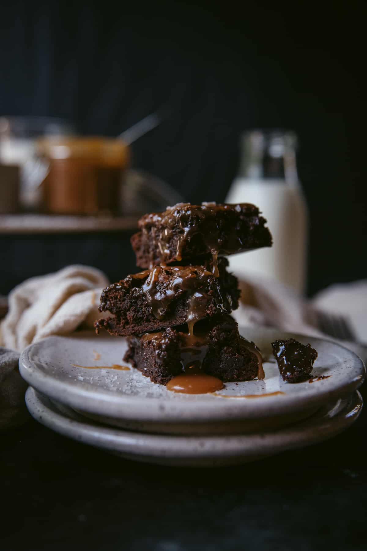 3 piece of salted caramel brownie are stacked on top of each other on  a white terrazzo plate. Caramel has ran over all the brownies and there is a small pool gathered on the plate. A piece of brownie has broken of one of the pieces and sit on the plate. There is a bottle of milk in the background along with a jar of the caramel sauce.
