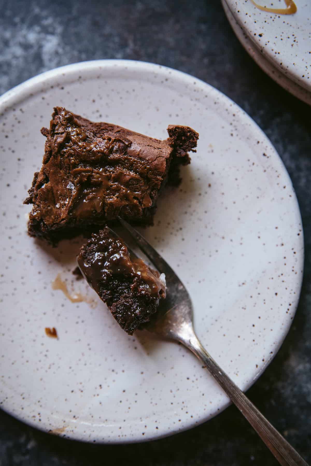 A piece of salted caramel brownie is sitting on a white terrazzo plate. A fork which has sliced into the brownie lays on the plate next to it.