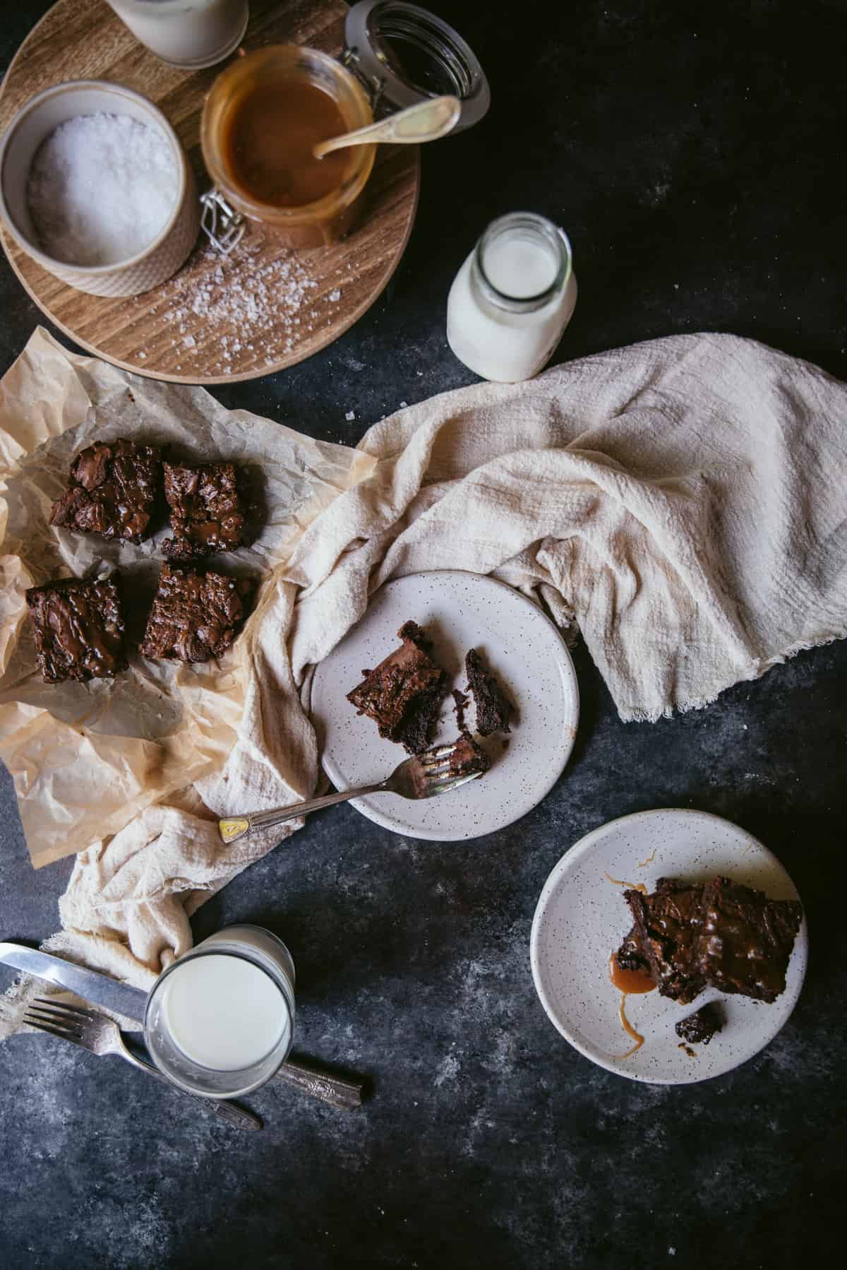 stack of 3 brownies on one plate and a single brownie on another speckled plate with a fork and bite taken out of the brownie.  Cream colored linen napkin in the background with cups of milk, parchment paper with more brownies on top and ramekins of salt flakes and salted caramel.