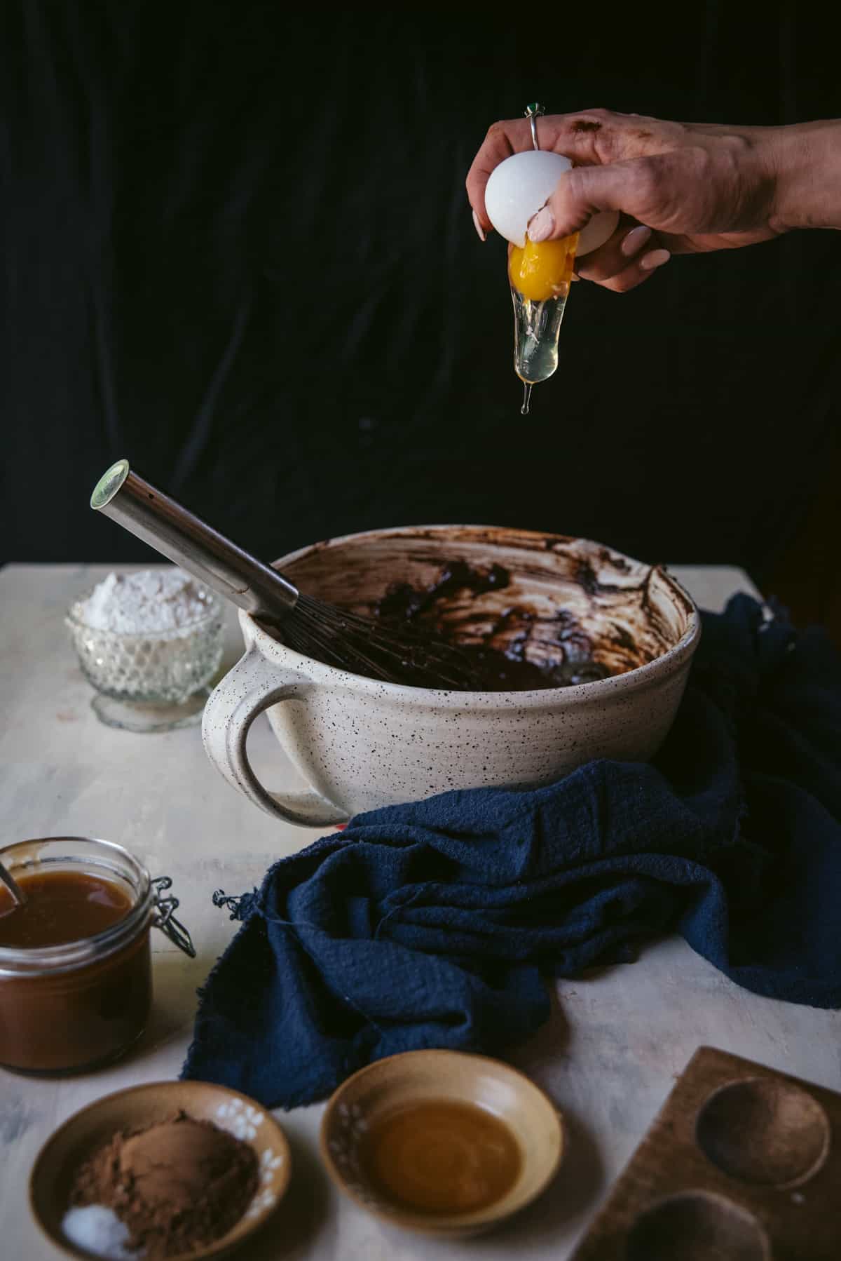 One hand cracking an egg into a ceramic speckled mixing bowl.  The egg is slowly falling from the egg shell.  On the table is a blue linen napkin and ramekins filled with ingredients.