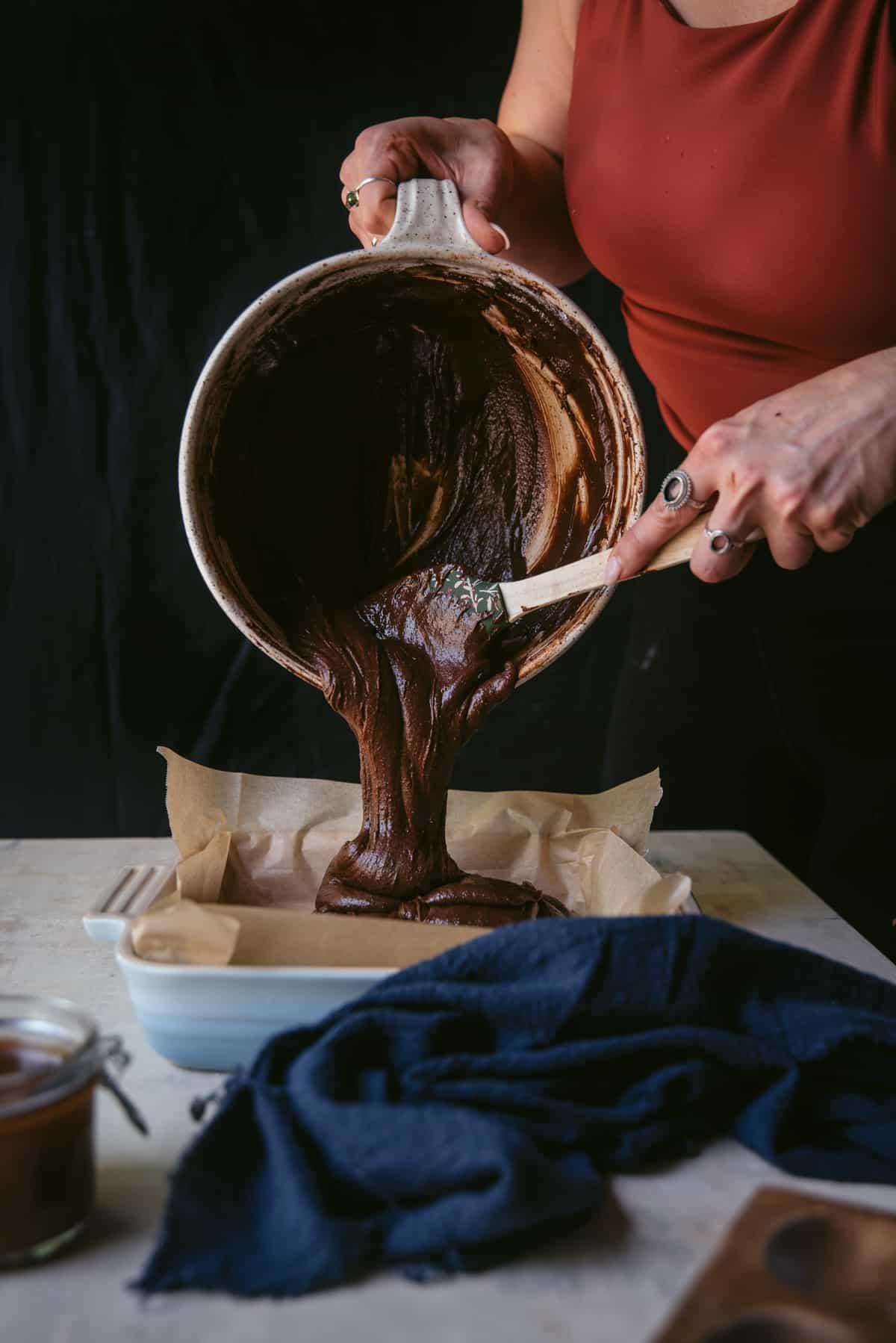 Straight on shot of brownie batter being scraped out of a mixing bowl into a blue 8x8 ceramic pan lined with parchment paper.  Blue linen napkin is on the table in front of the pan.