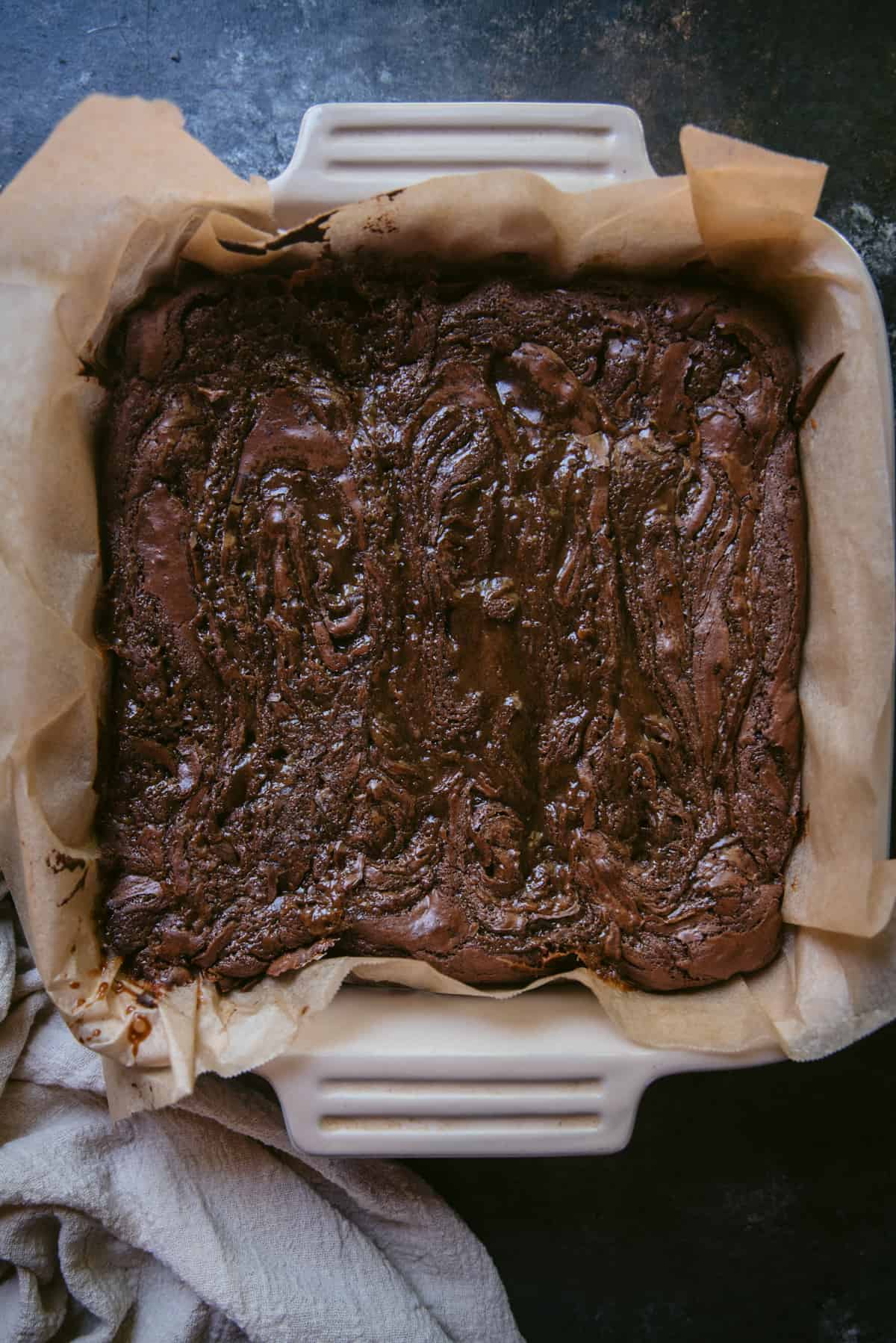 Overhead image of baked brownies in a ceramic 8x8 pan lined with parchment paper.  The brownies are crispy on top with swirls of salted caramel.