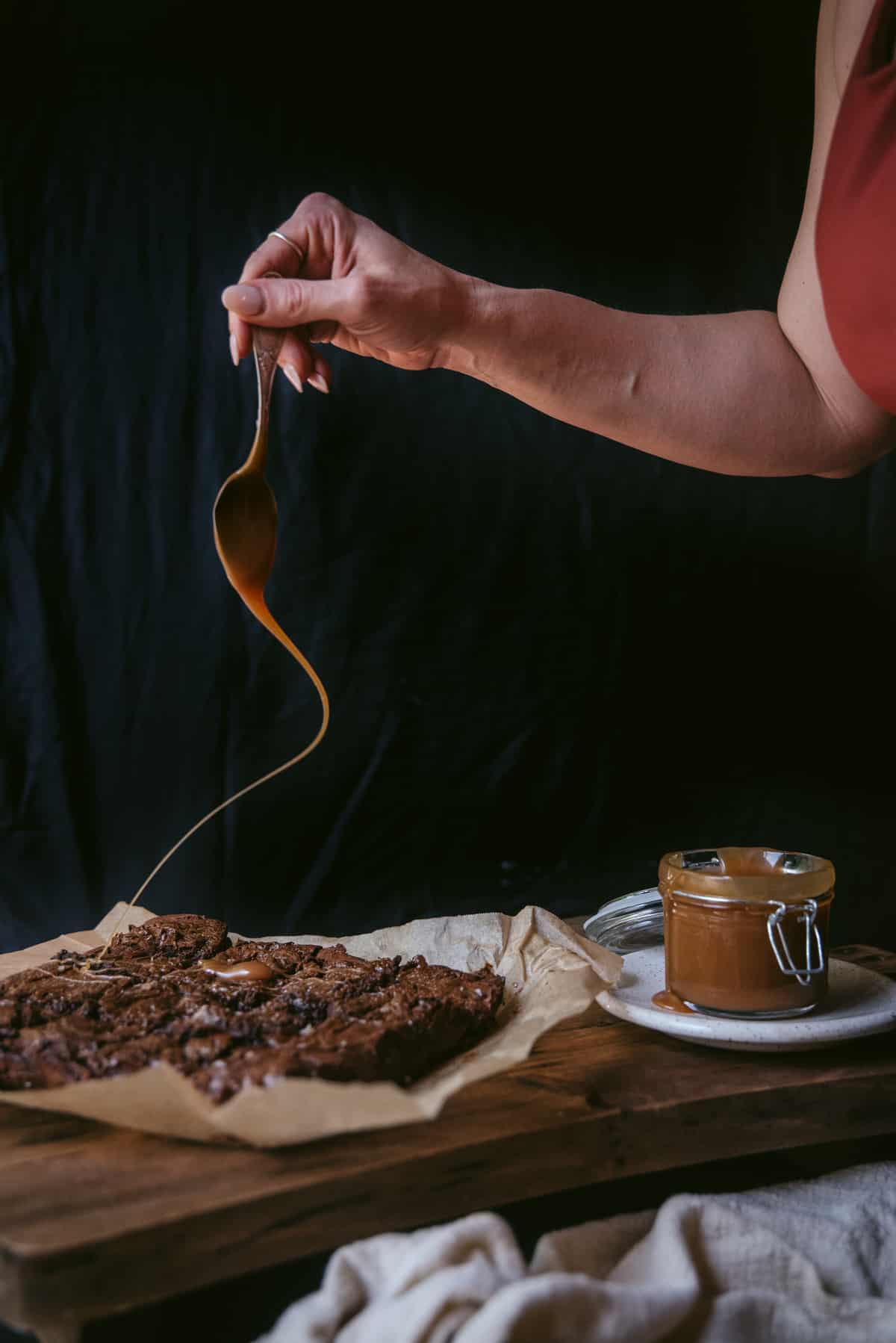 The brownies have been baked and sliced but remain on the baking paper on top of a wooden table. The jar of salted caramel is placed on a white plate next to it. A hand holding a spoon is drizzling caramel all over the top of the brownies.