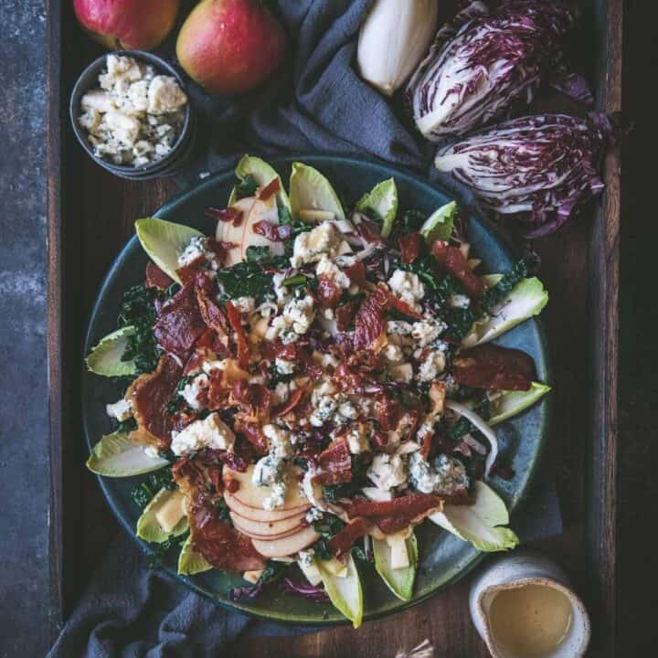 overhead image of salad on a large platter surrounded by whole ingredients. On the platter there is sliced kale, apples, crispy pieces of prosciutto and crumbled blue cheese on top surrounded by leaves of endive fanned along the outside of the plate.