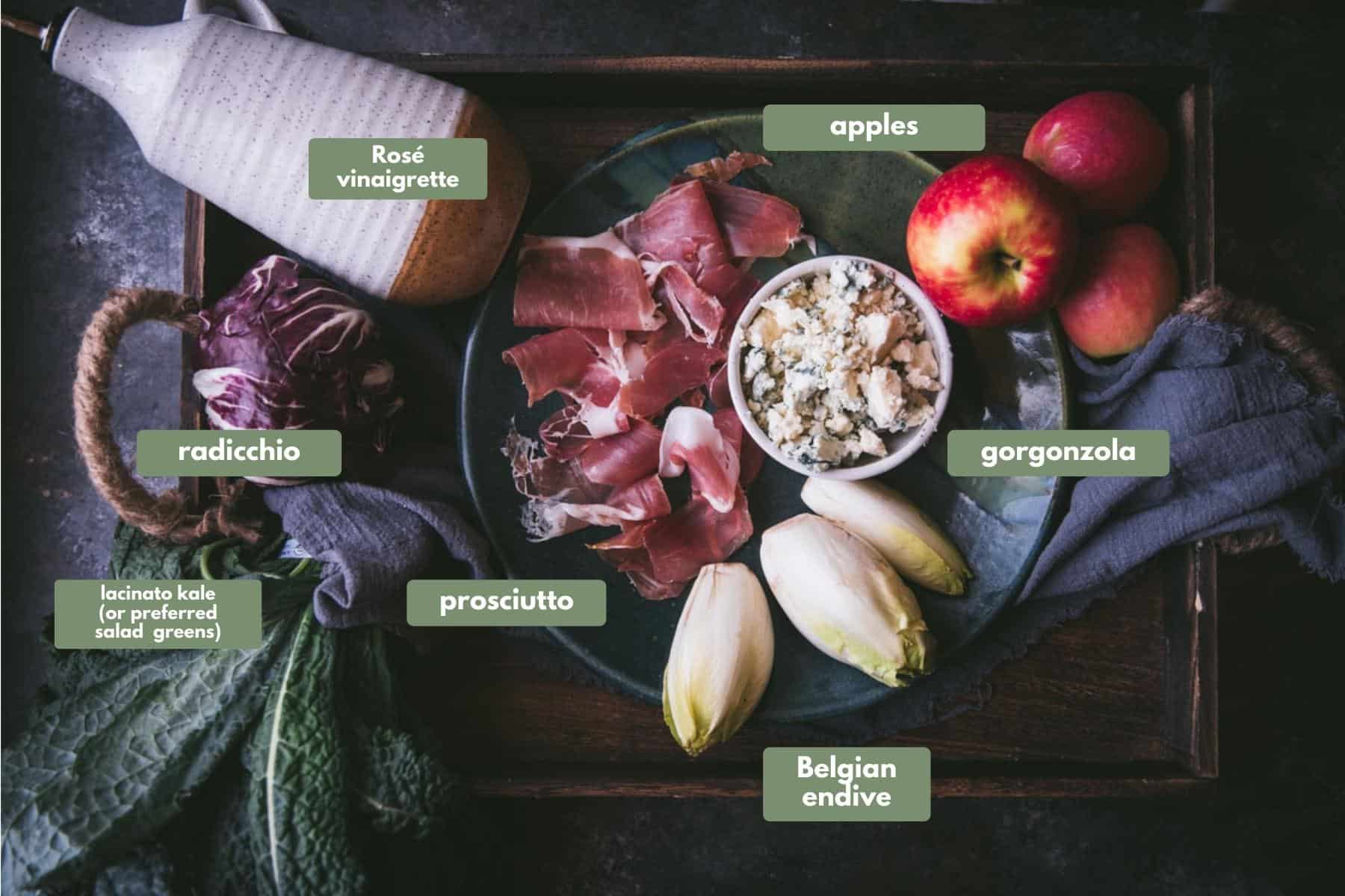 salad ingredients on a ceramic platter over a linen napkin tucked inside a wooden tray.  Ingredients include kale, endive, radicchio, apples, prosciutto, gorgonzola and a ceramic pour bottle of dressing.