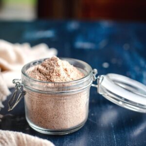 close up photo of everything but the elote seasoning in a glass jar with a lid open on top of a blue background next to a cream colored linen napkin.