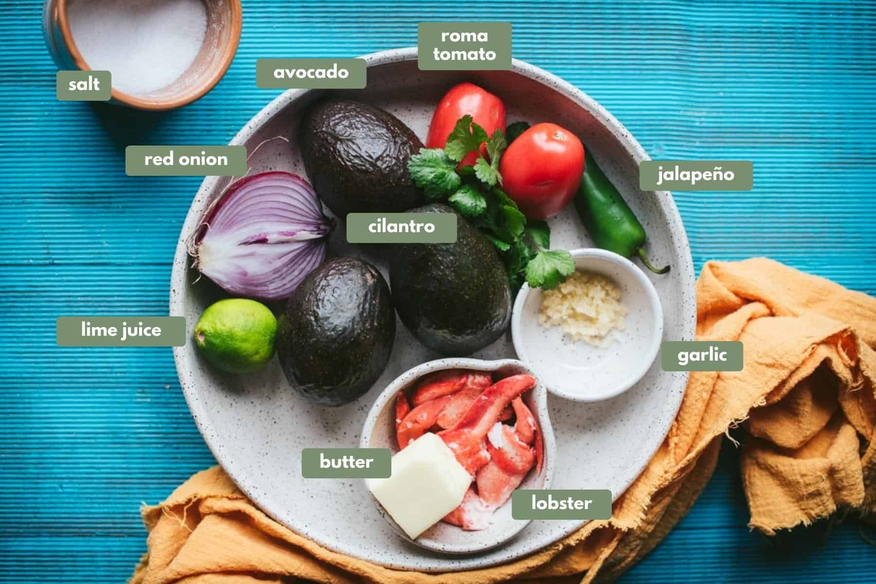 Ingredients for lobster guacamole on a white terrazzo plate, on a blue table cloth. 
All the ingredients on the plate are; half a red onion with the middle side showing up, 3 avocados,  2 tomatoes, a jalapeno, small hand full of cilantro, garlic in a ramekin dish, lobster tails with a chunk of butter on top in another ramekin dish. The salt is in another small bowl next to the plate of ingredients.