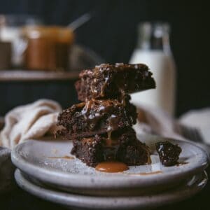 3 piece of salted caramel brownie are stacked on top of each other on a white terrazzo plate. Caramel has ran over all the brownies and there is a small pool gathered on the plate. A piece of brownie has broken of one of the pieces and sit on the plate. There is a bottle of milk in the background along with a jar of the caramel sauce.