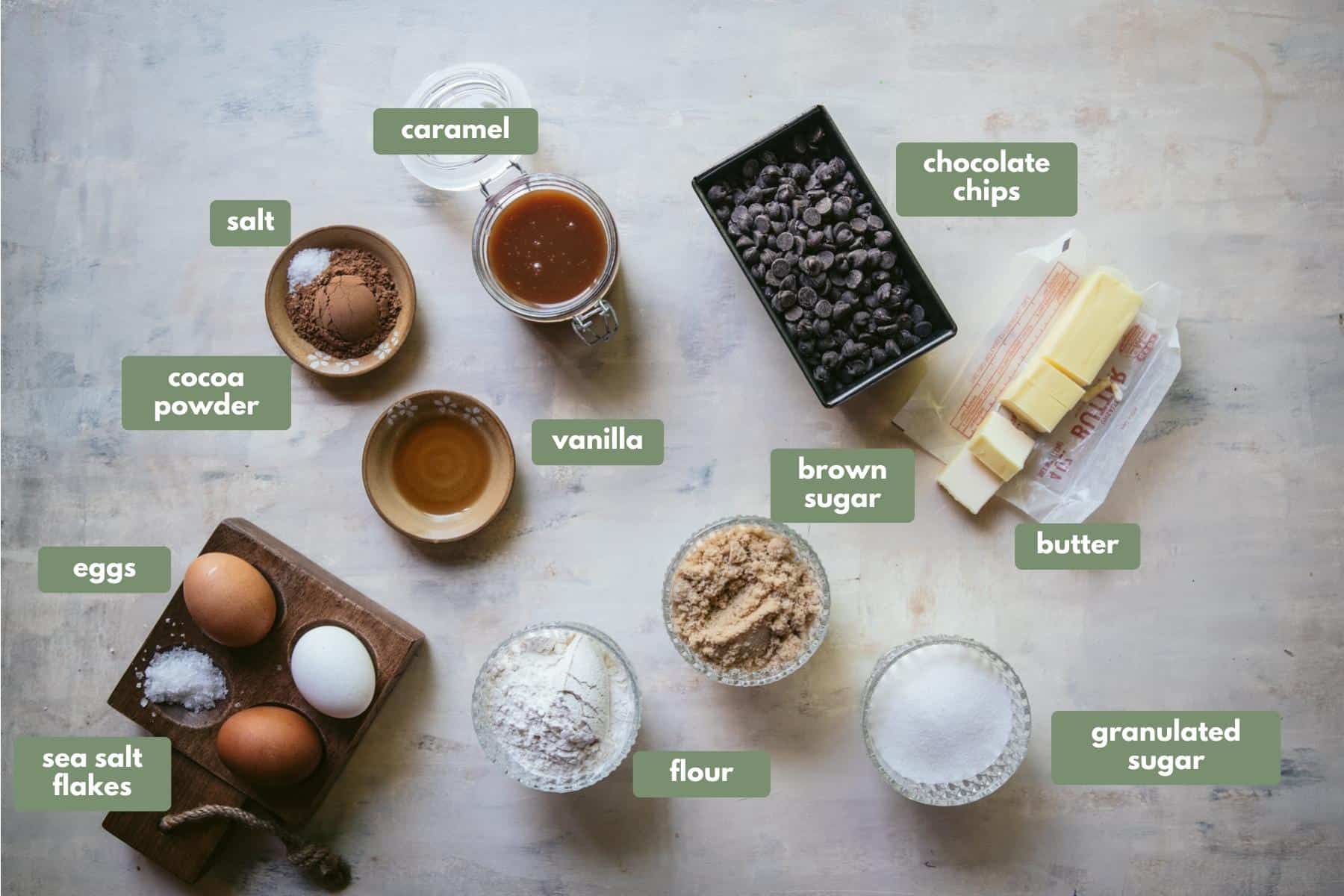 All of the ingredients used in the recipe placed in separate containers sitting on a white countertop. 
Moving clockwise there is a glass jar of caramel, a tub of chocolate chips, butter sitting on top of the wrapping, granulated sugar, brown sugar and flour all sitting in they're own glass bowls, 3 eggs in a wooden egg holder, in the 4th egg compartment there is sea salt flakes. Then there is vanilla in a brown ceramic dish, followed lastly by cocoa powder and sea salt together in another brown ceramic dish.