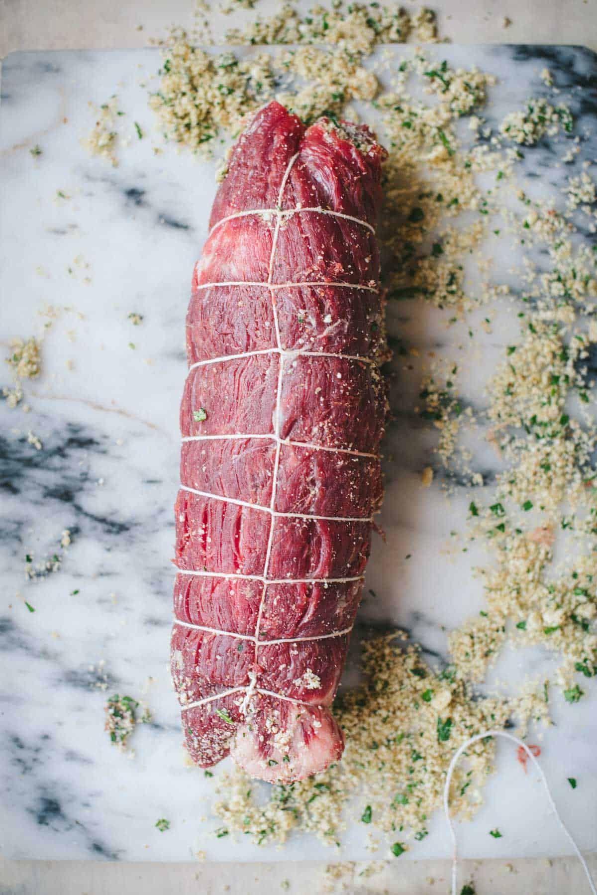 The braciole roll is sitting on a white marble chopping board and string has been tied around it to keep it from falling apart.