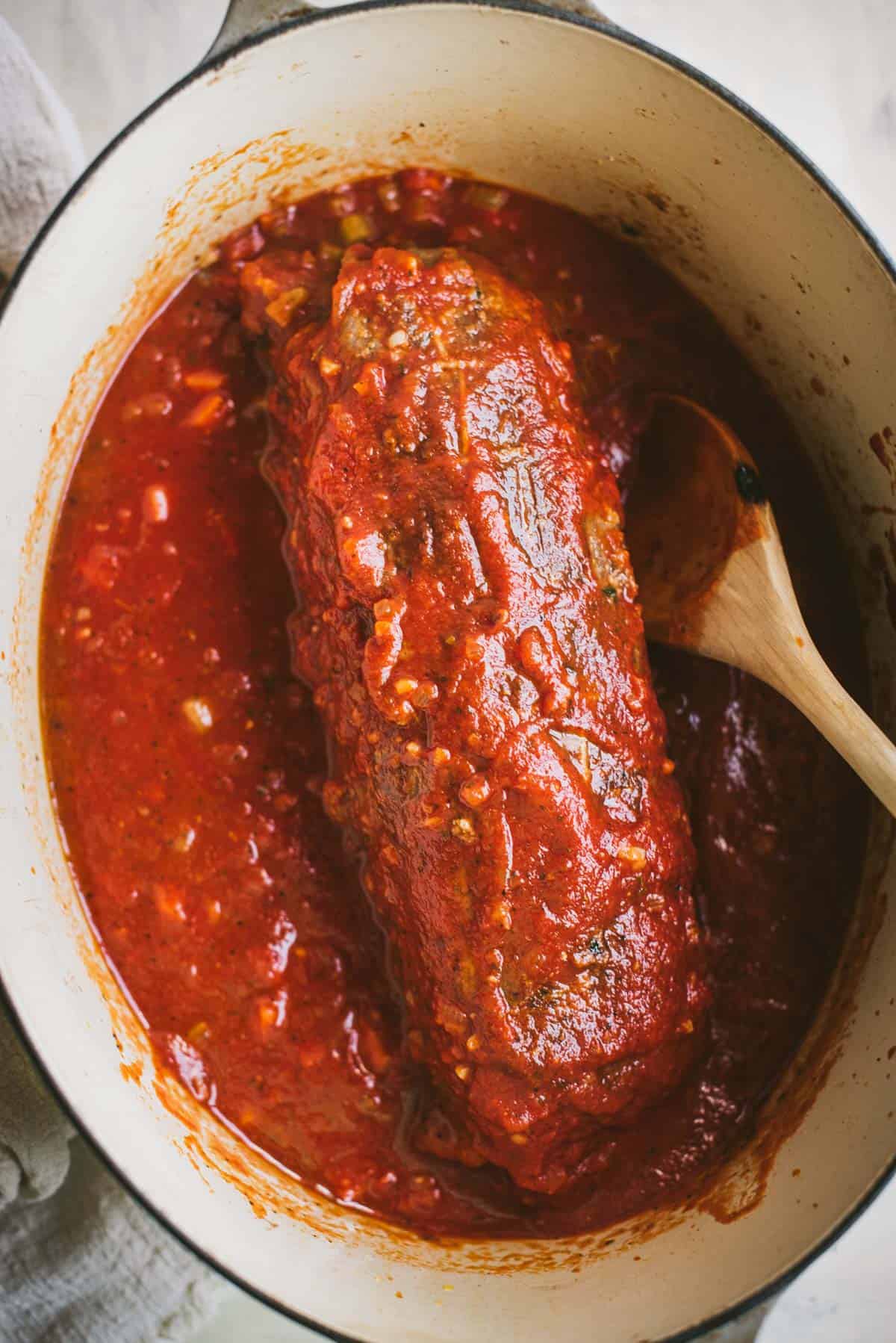 Tomato sauce has been added to the large white pot. The tomato is covering the braciole roll and has been mixed into the wine mixture.