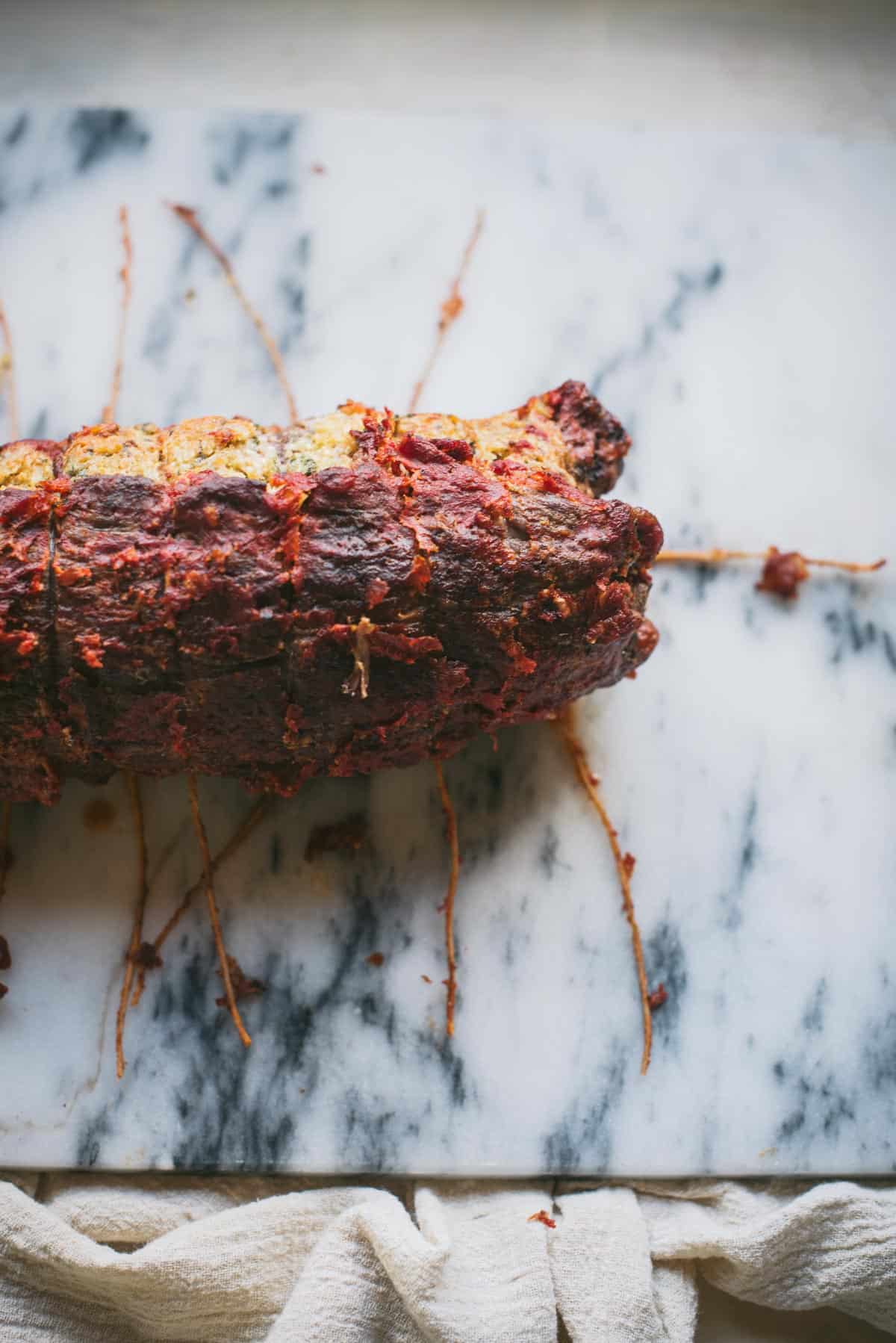 The cooked beef braciole is sitting in a white marble chopping board. The strings have been cut and the meat is resting.