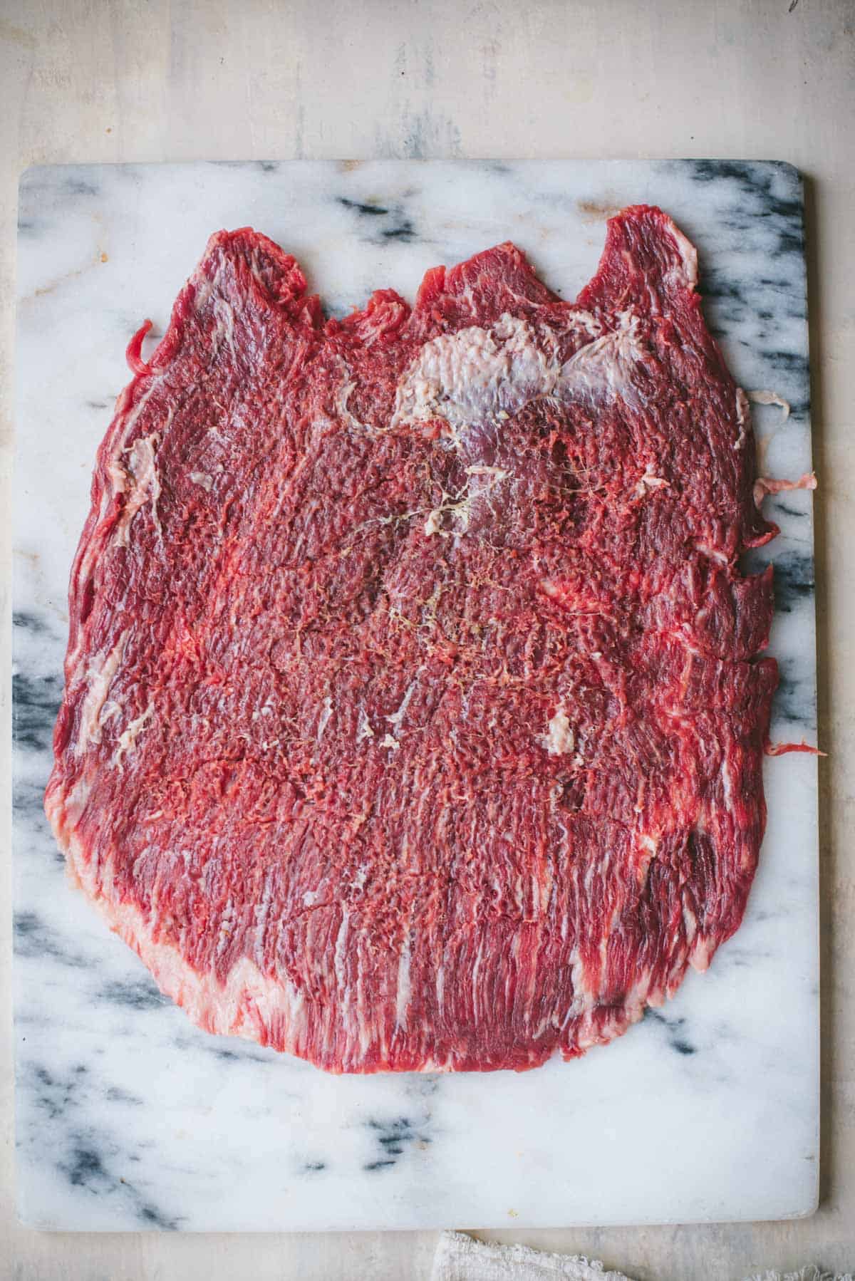 A large piece of flank steak is resting on a white marble chopping board on a white counter. 