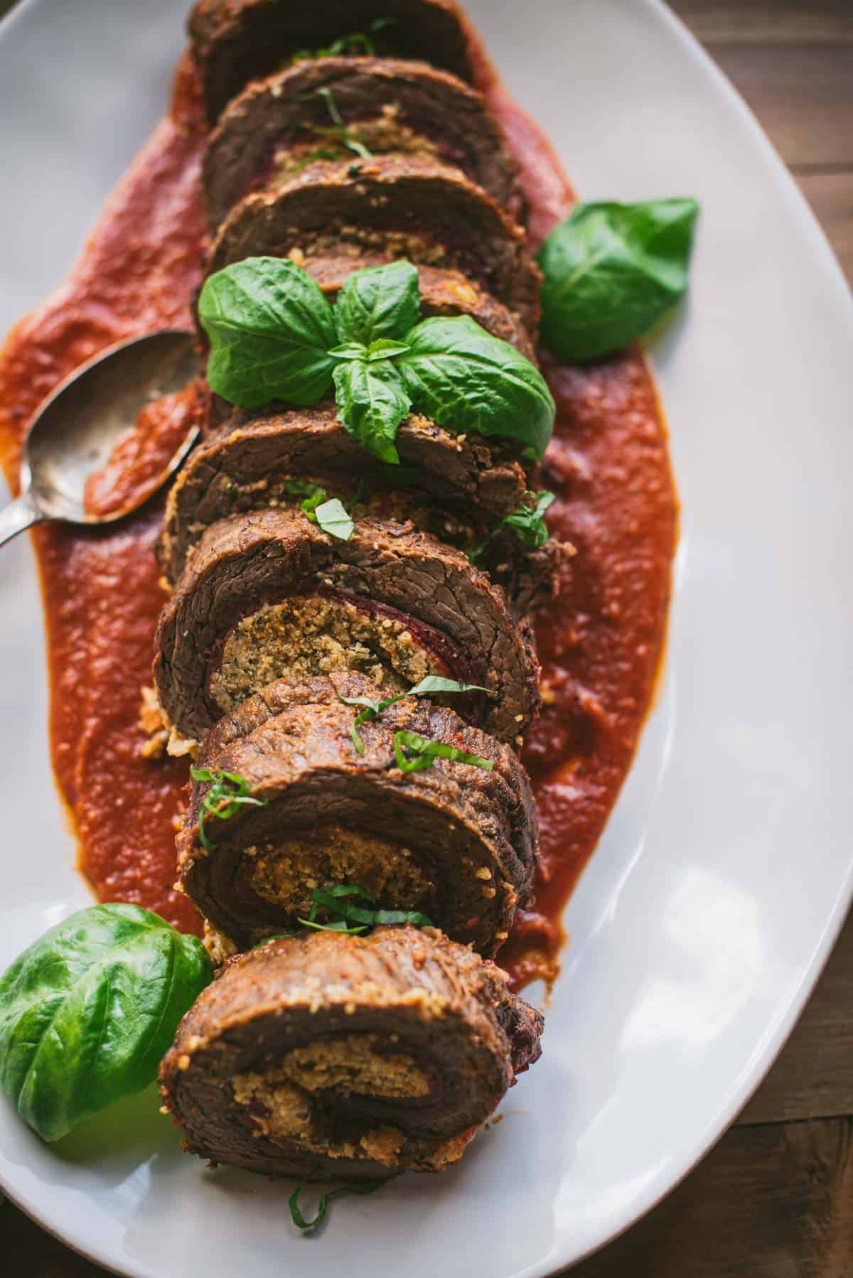 Beef Braciole sliced up and placed on top of a tomato sauce on a white plate. It is garnished with fresh basil leaves.