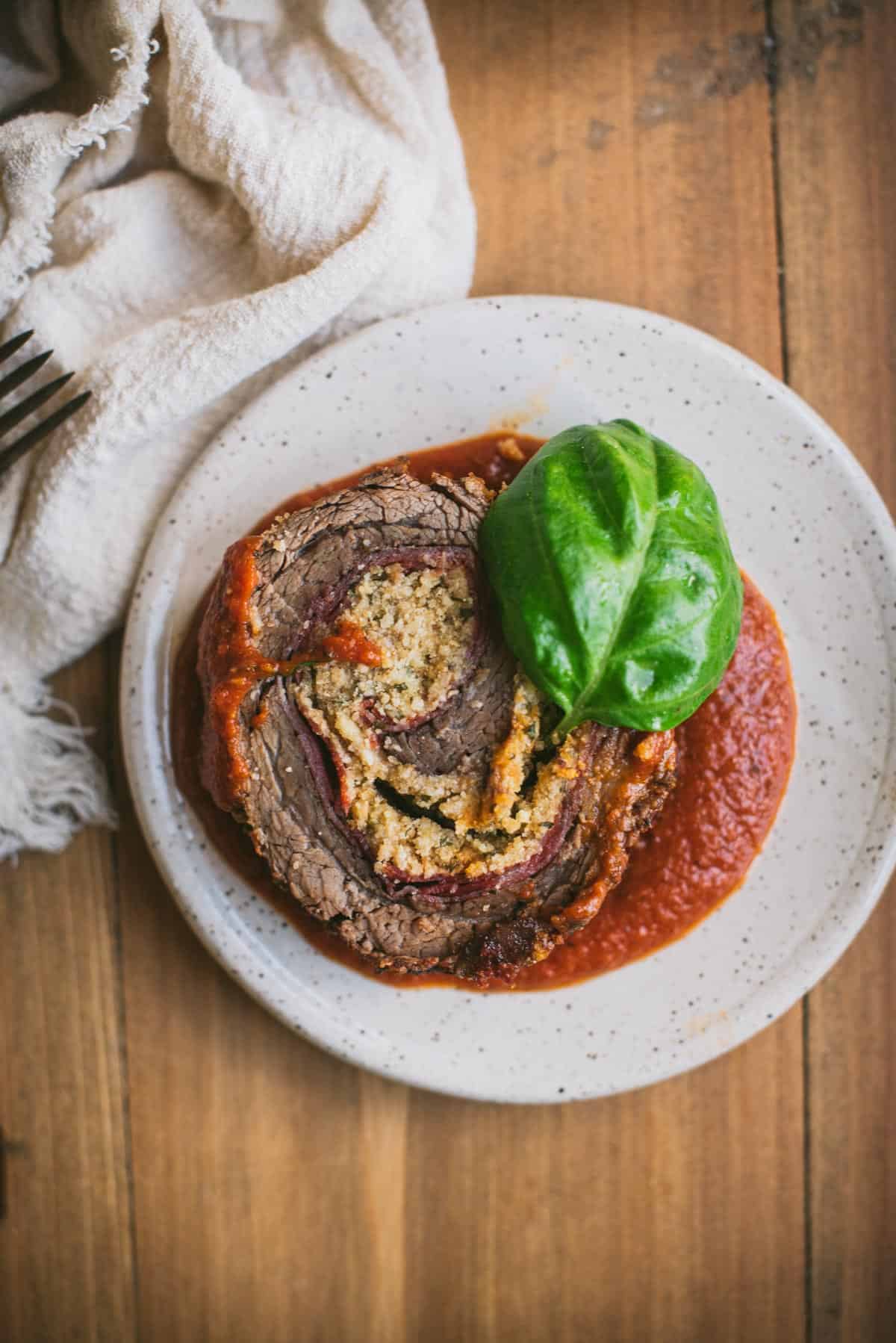 1 piece of beef braciole on a white terrazzo plate on a wooden table. The meat is sitting on a small amount of tomato sauce and a basil leave is sitting on top of the swirled meat and cheese.