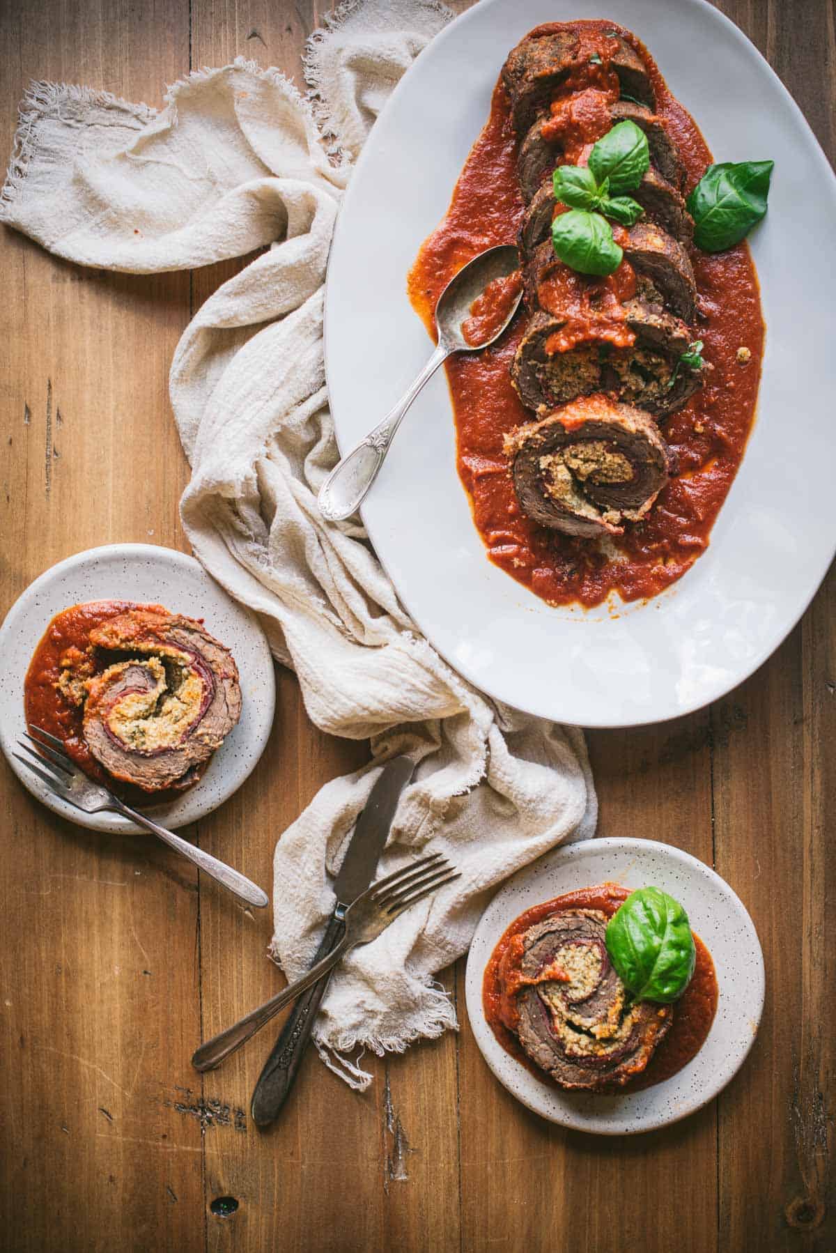 Italian Beef braciole on a bed of tomato sauce on a white plate. The braciole is sliced into pieces and topped with fresh herbs.
There are 2 smaller white plates on top of the wooden table. Each plate has a slice of braciole and tomato sauce on top. There is some discarded cutlery sitting on the table too.