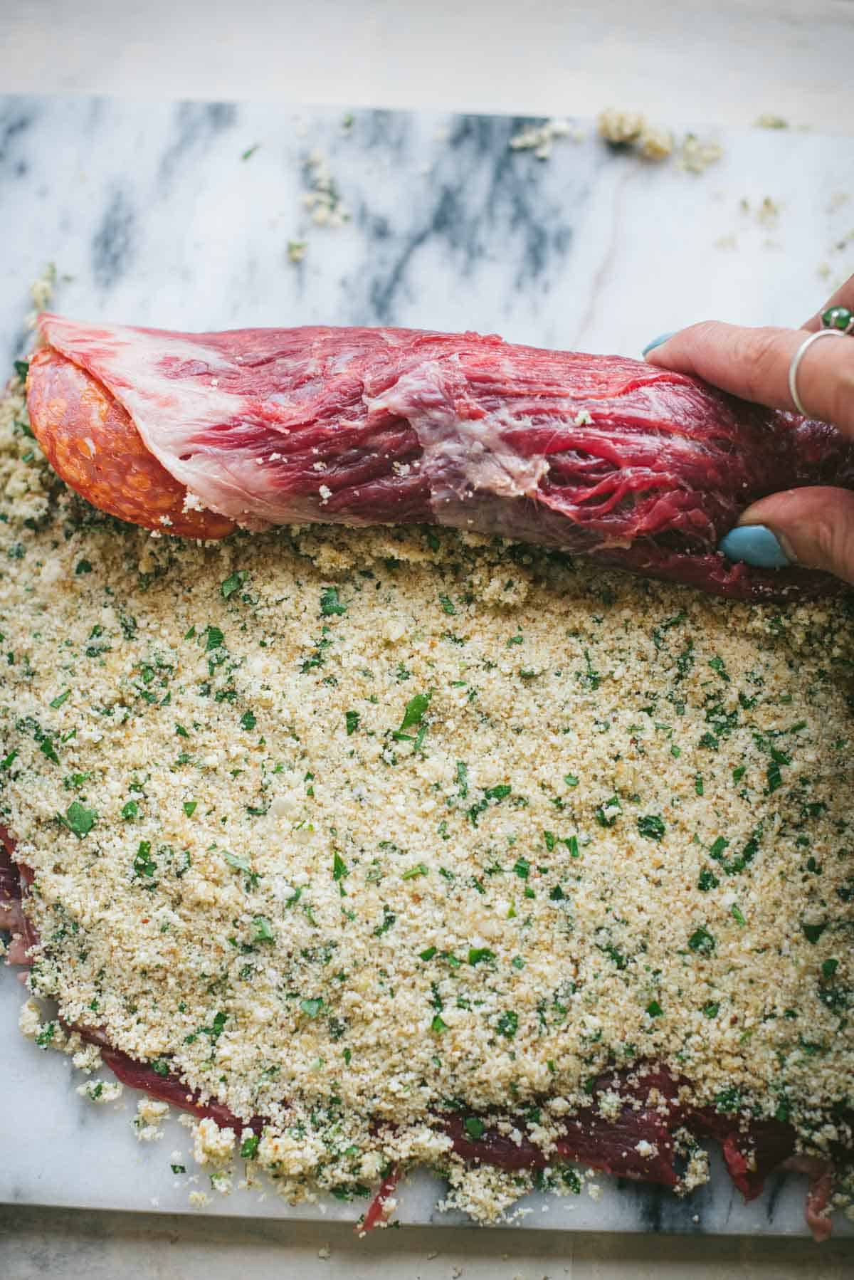 The flank steak has now had the cheese and breadcrumb mixture layered on top of the cured meats. There is a hand rolling the meat up into a log - like a cinnamon roll.