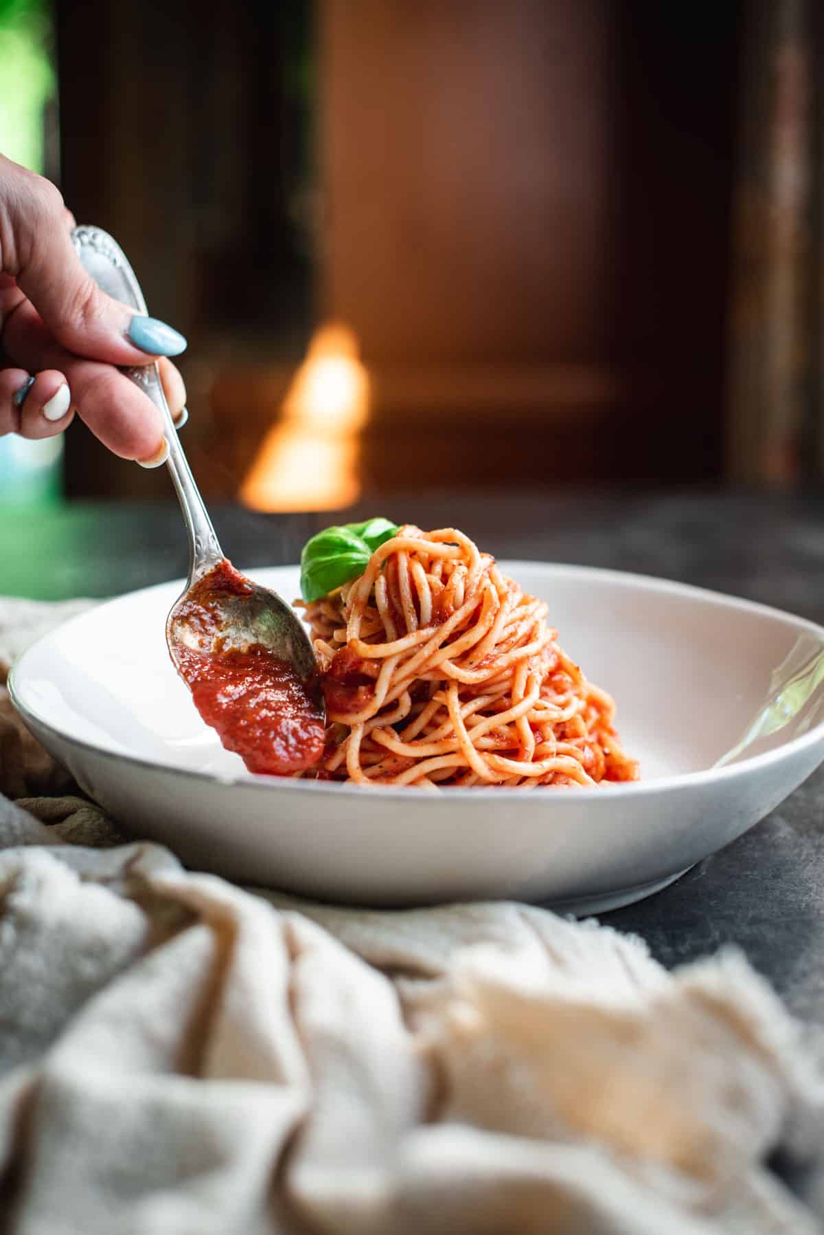 A hand holding a silver spoon is placing  easy homemade pasta sauce over spaghetti in a white bowl. It is garnished with a basil leaf and sitting on a grey counter.