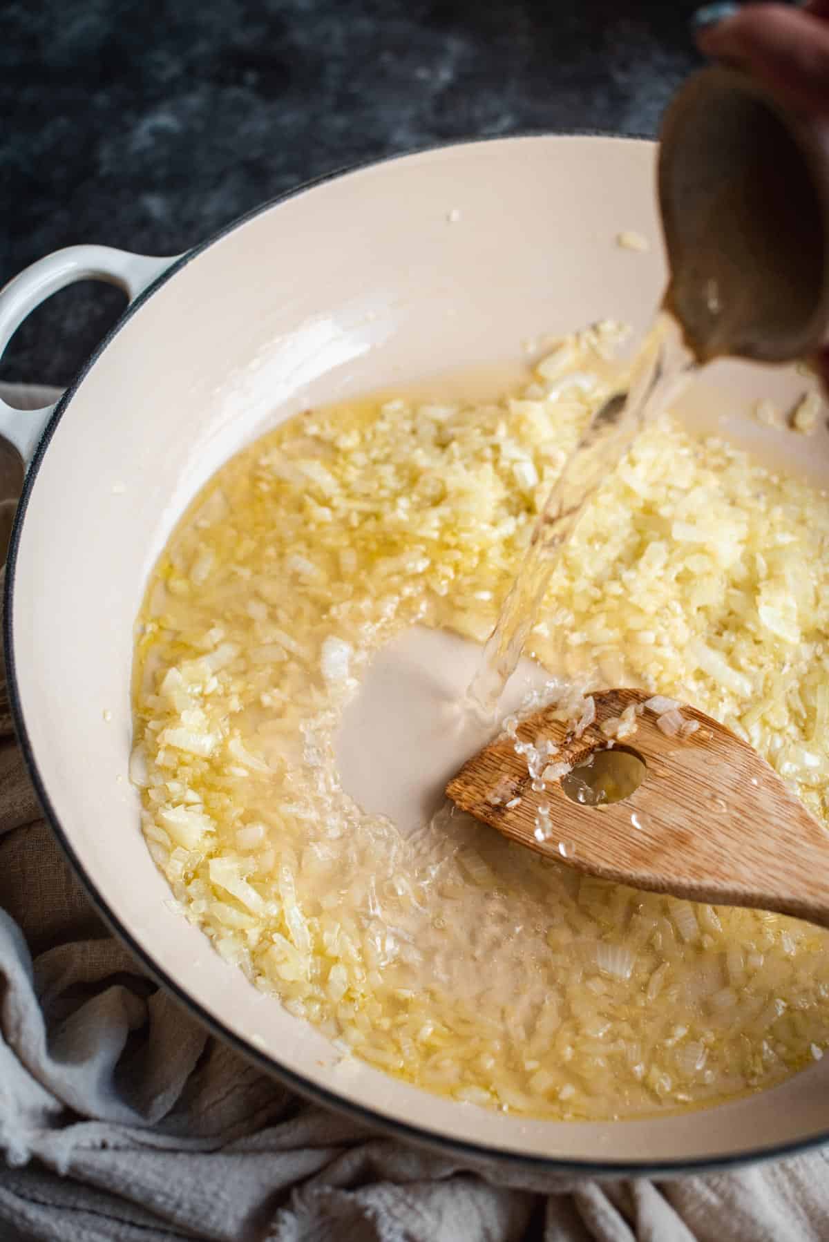 A jug of white wine is being poured into a white saucepan, to cover the cooked onion and garlic, and is stirred with a wooden spoon.