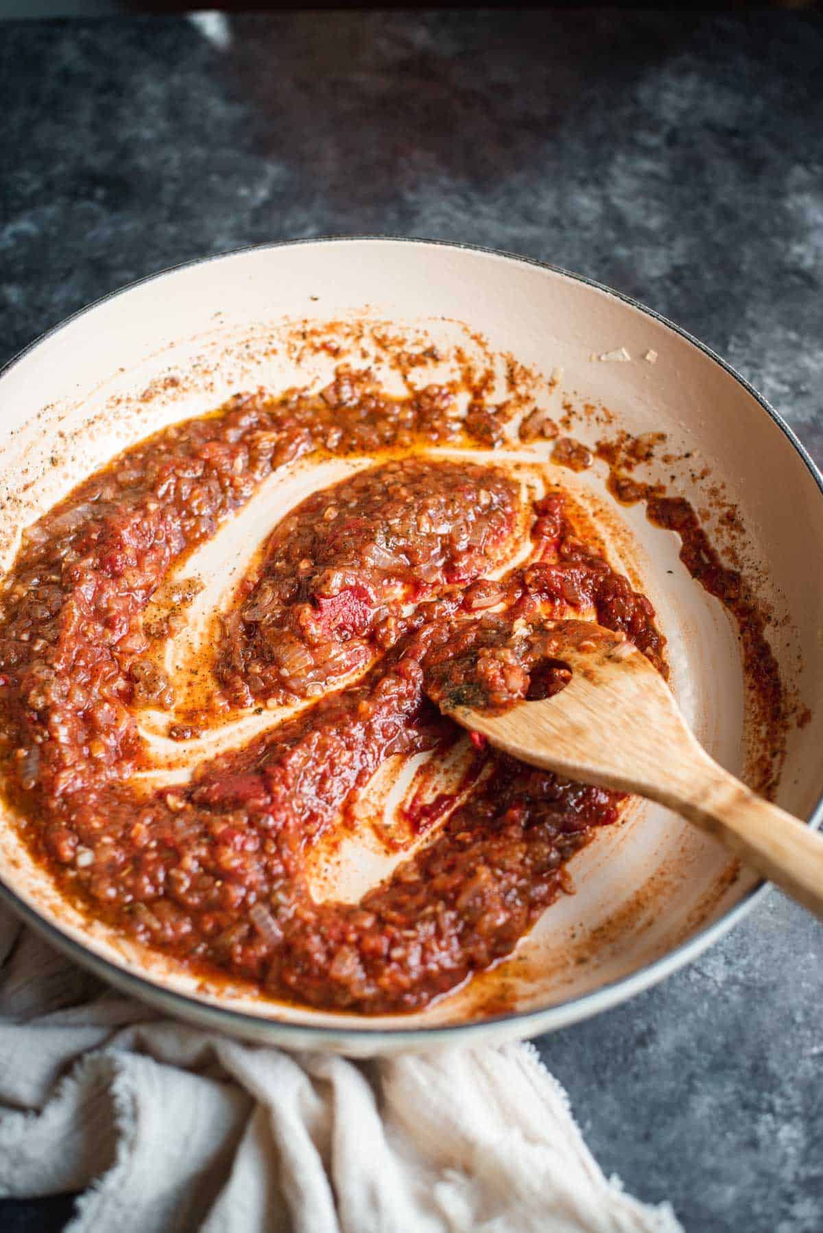 Tomato paste has been added to the mixture so the sauce is now a brown/red color. It looks very chunky and thick. A wooden spoon is stirring the mixture.