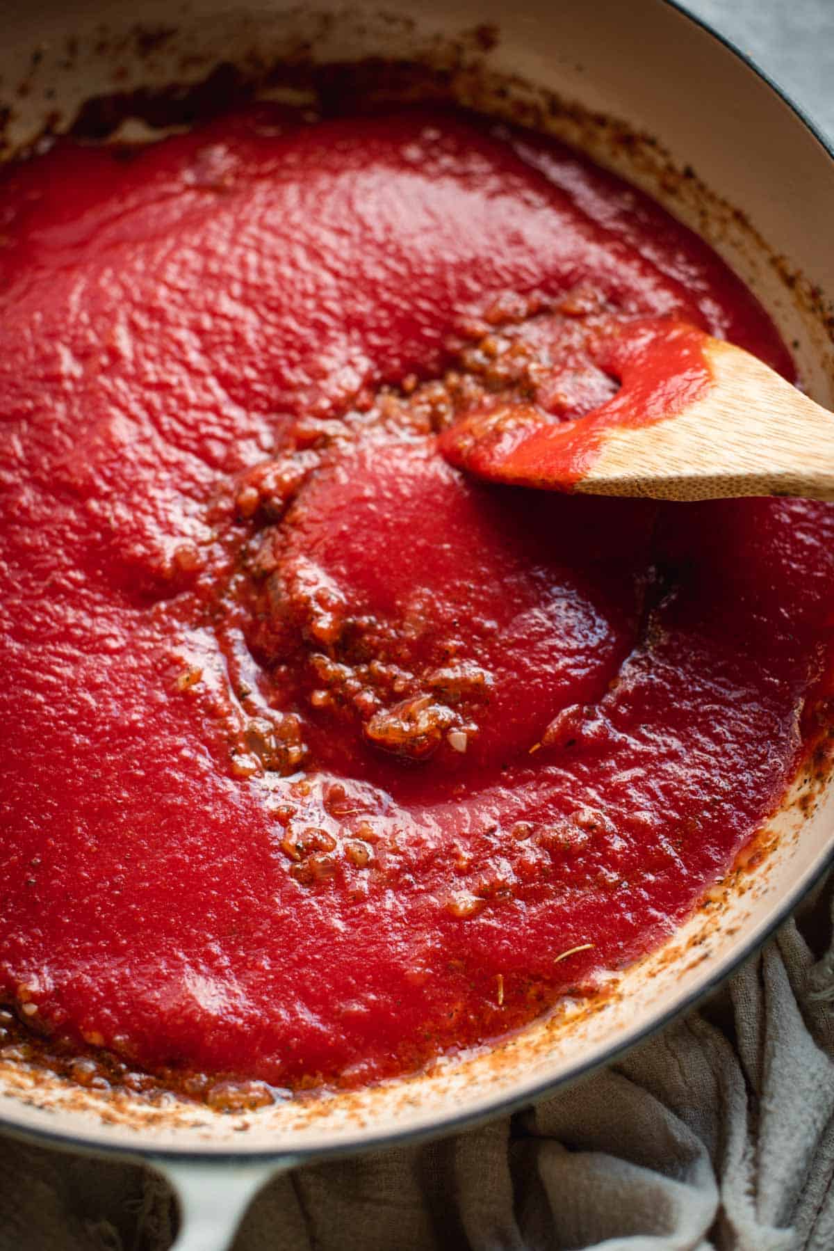 Canned tomatoes are added on top of the sauce in the white saucepan. A wooden spoon is about to incorporate all the ingredients together.