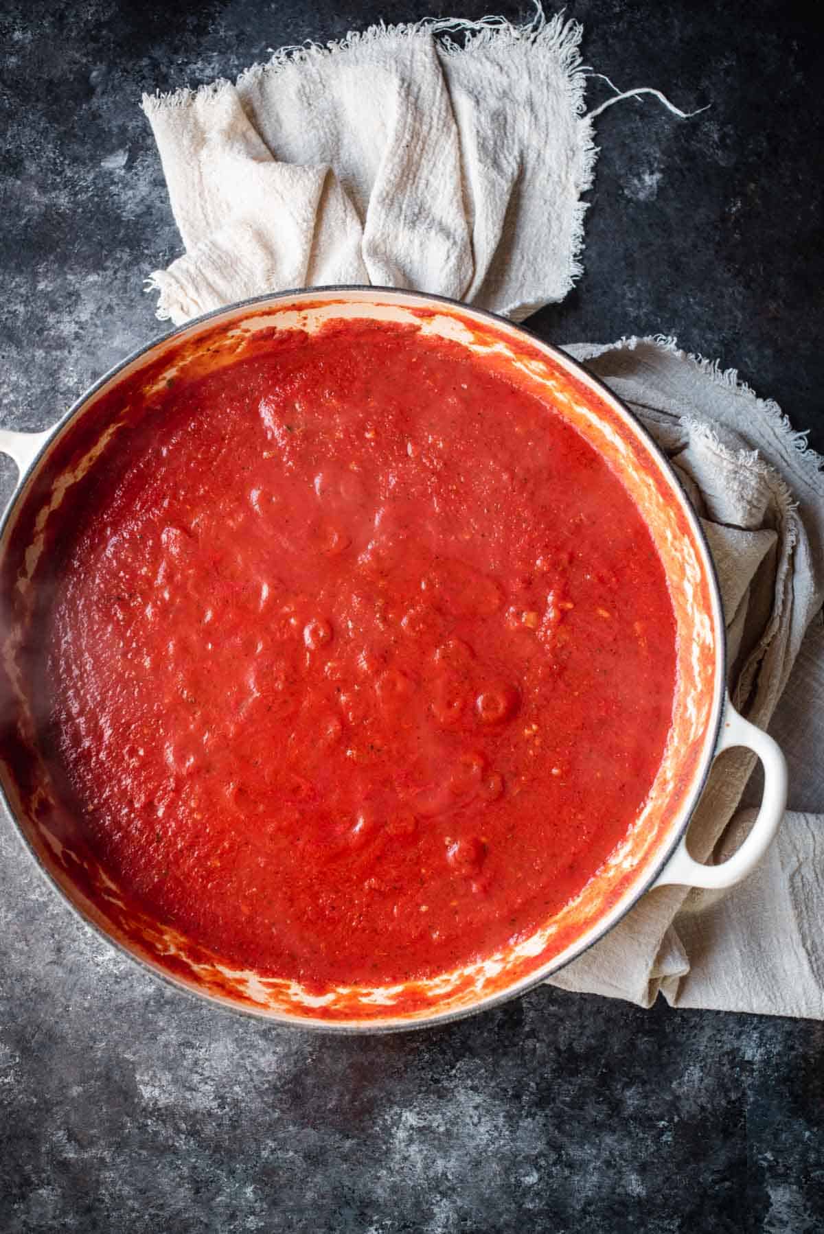 A slate grey counter top has a cream cloth laying on it and a big white saucepan filled with easy to make pasta sauce. The fresh pasta sauce is bright red in color and looks like it's still very hot.