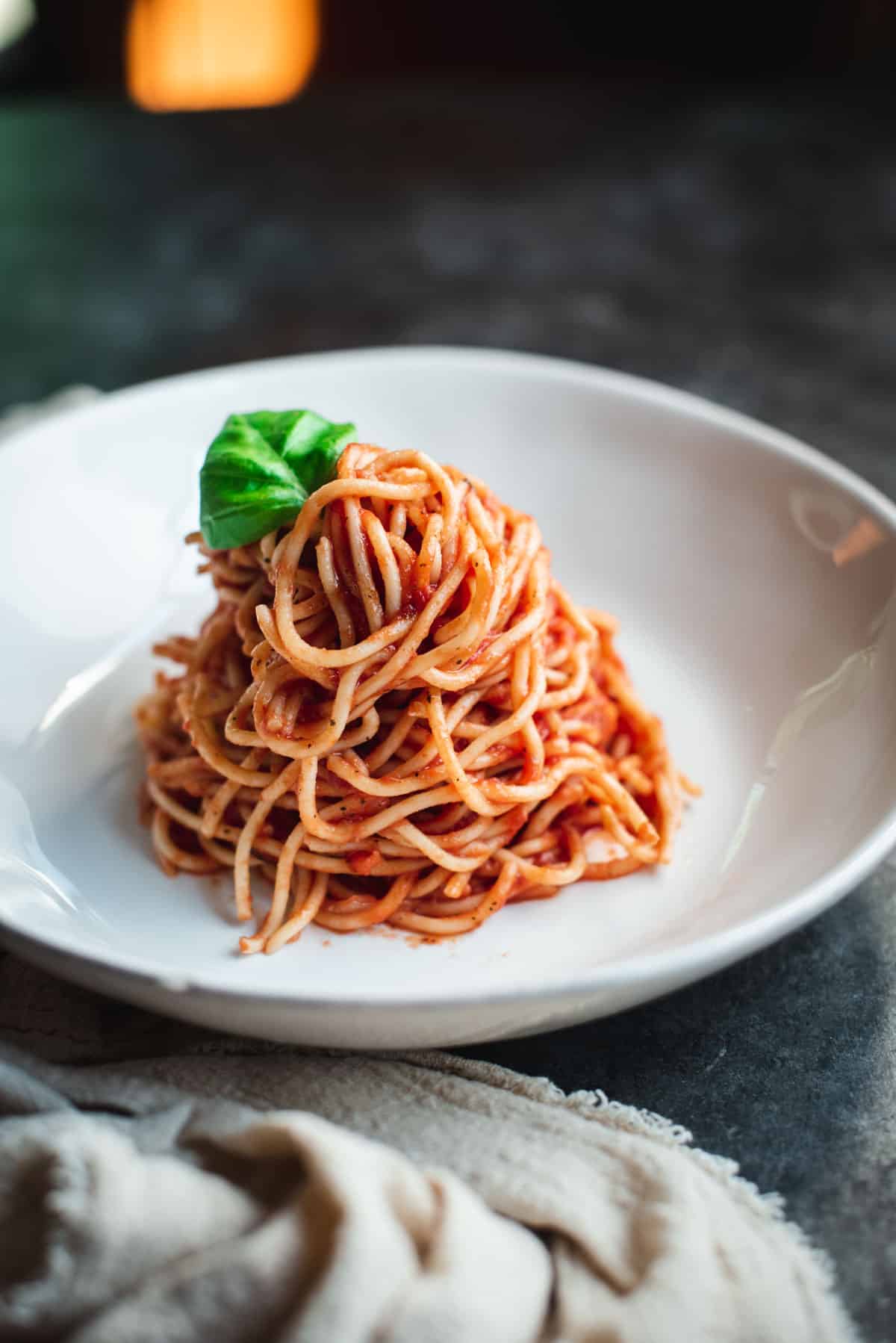 A tower of spaghetti mixed with the homemade pasta sauce is in a white bowl and garnished with a fresh basil leaf.
