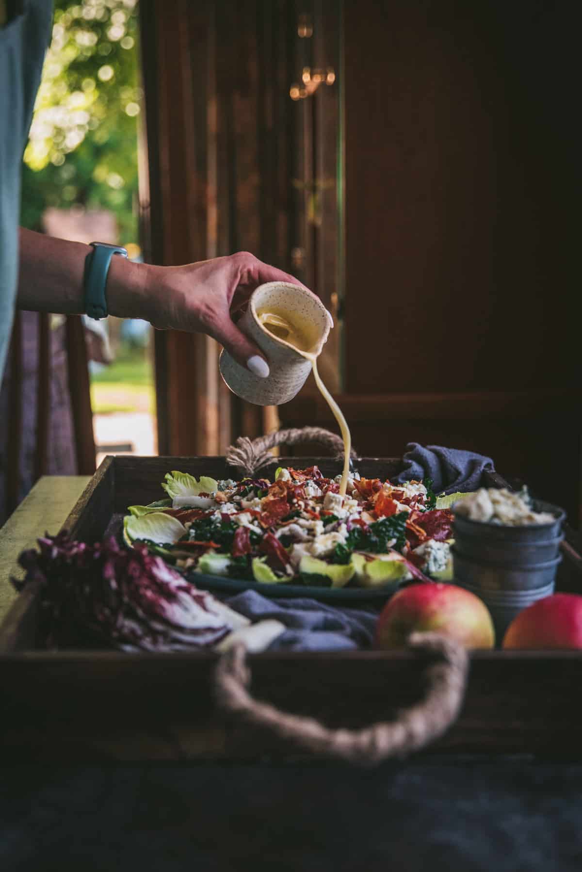 dressing being poured over a salad.  Wooden hutch and green trees through a window are in the background.