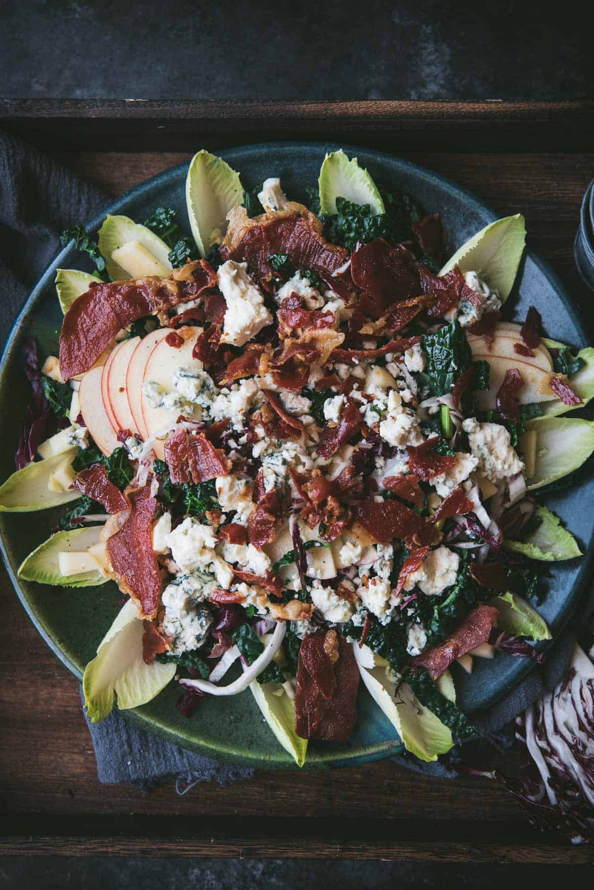 overhead close up of endive radicchio salad with kale, apples, gorgonzola crumbles and crispy prosciutto on top.  All the ingredients are layered on a green and blue platter inside of a wooden serving tray.