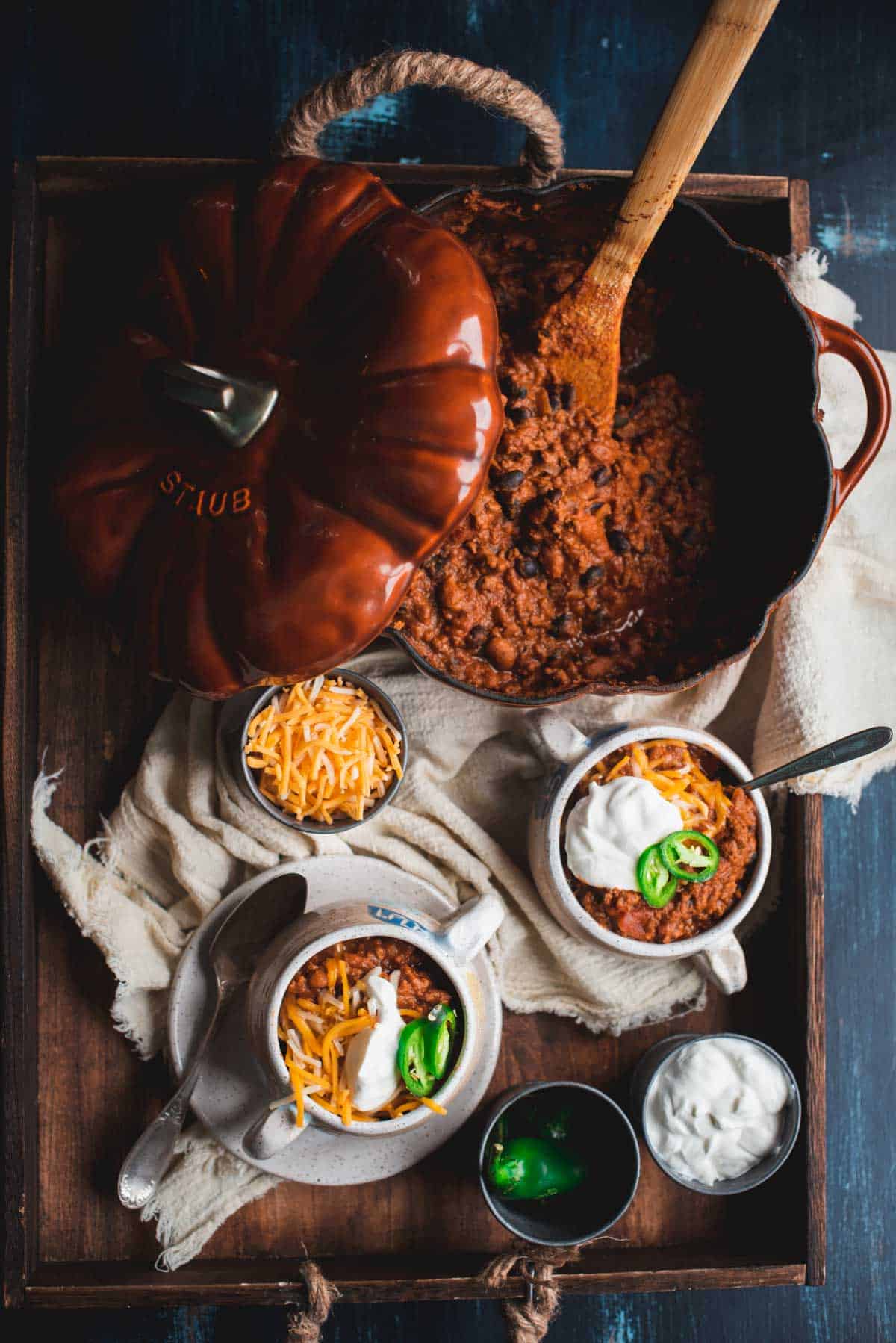 A saucepan in the shape and color of a pumpkin sits on a wooden tray and is filled with Pumpkin chili, the lid is leaning on the edge of the pan. There is a small bowl of grated cheese on the board, alongside 2 white bowls of Pumpkin Beef Chili. There is extra jalapenos and sour cream in ramekin dishes to the side.