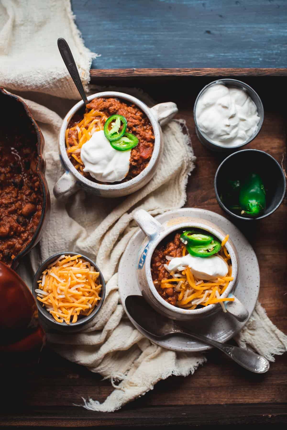 2 white bowls of pumpkin beef chili sitting on a tablecloth on a wooden tray.  The chili is topped with cheese, sour cream and jalapenos. There is a bowl of grated cheese on the tray, also sour cream and a pot of jalapenos. Part of the pumpkin shaped pot with the chili inside can be seen also.