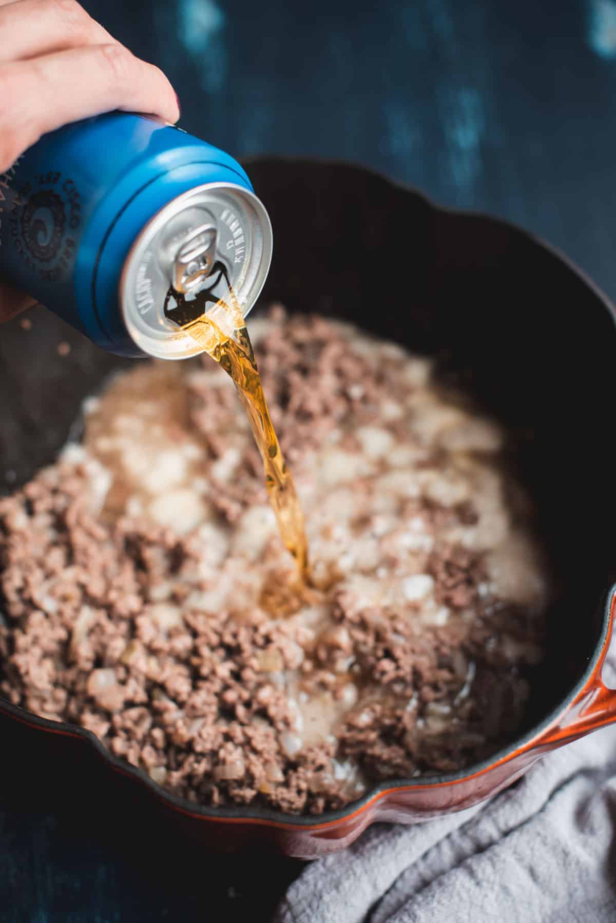 A can of beer is being poured into the beef and onion mixture sitting inside the pumpkin shaped pot on the burner.