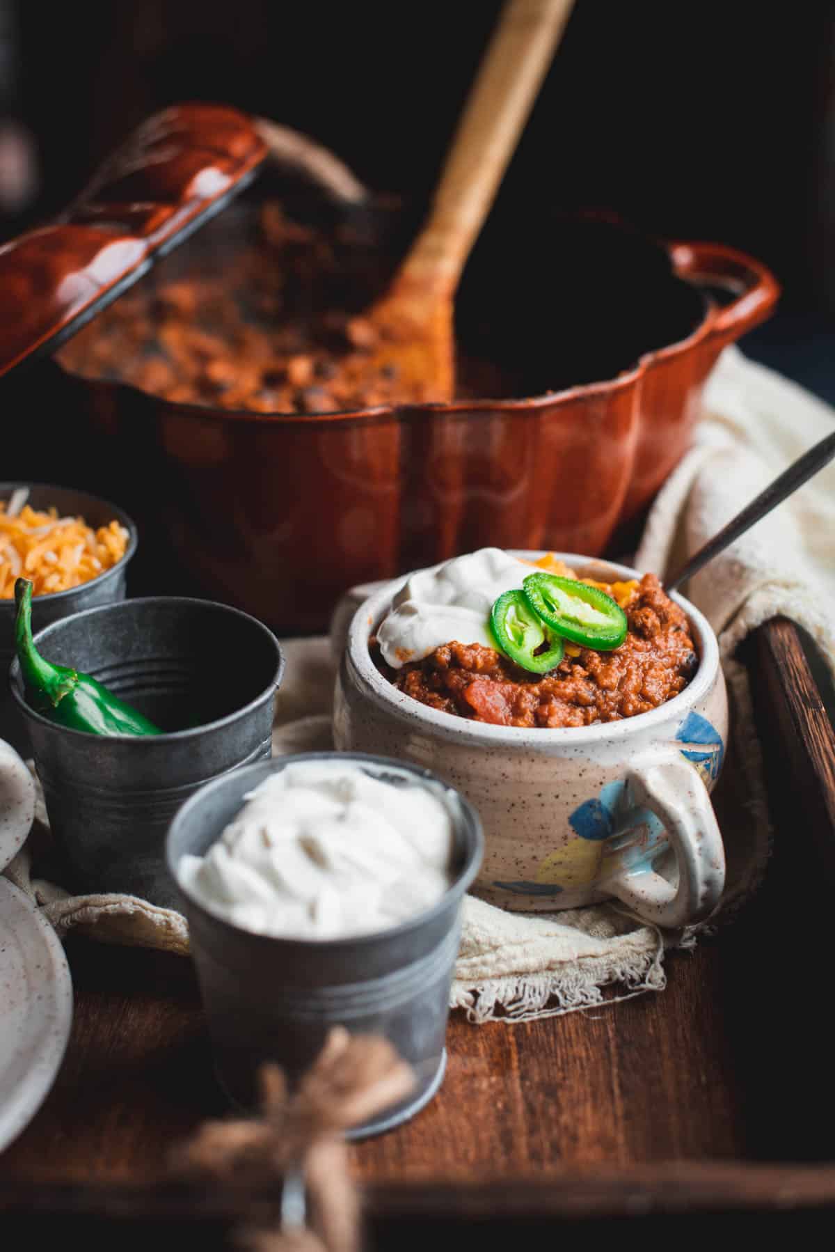 In the background is a pumpkin shaped pot containing chili and a wooden spoon. In the foreground is a white bowl filled with pumpkin beef chili and topped with sour cream and jalapenos. There are small buckets next to it with grated cheese, jalapenos and sour cream.
