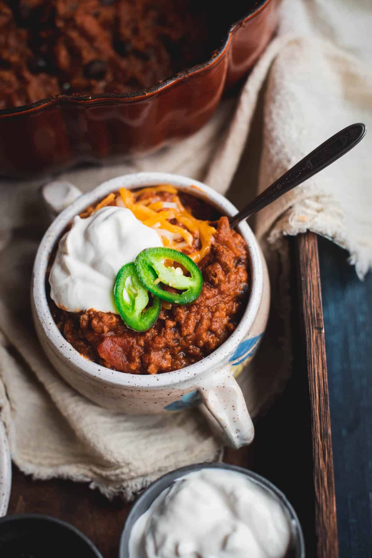 1 white bowl of pumpkin beef chili sitting on a tablecloth on a wooden tray.  The chili is topped with cheese, sour cream and jalapenos. In each corner, part of the chili pot can be seen and part of the pot containing sour cream.