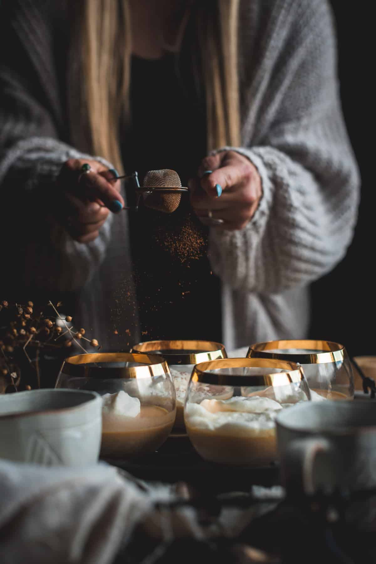 4 gold rimmed glasses are sitting on a crowded counter alongside other cups, dried flowers and  a cloth. A women is gently shaking cinnamon from a small sieve on top of the panna cotta desserts.