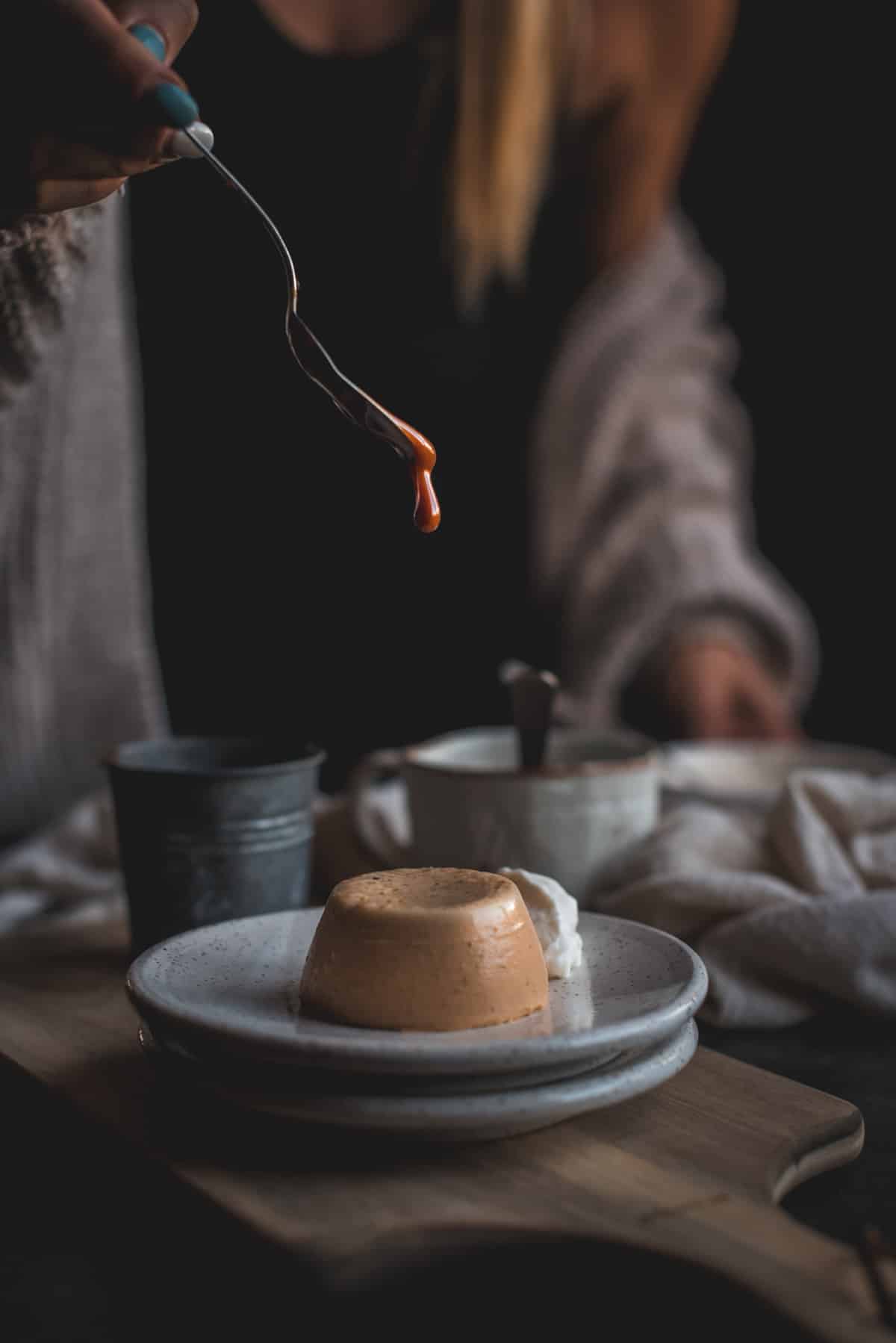 In a dark room, a woman wearing a chunky grey kintted cardigan is drizzling caramel on top of the set Pumpkin Panna Cotta which is sitting on a grey terrazzo  plate.
