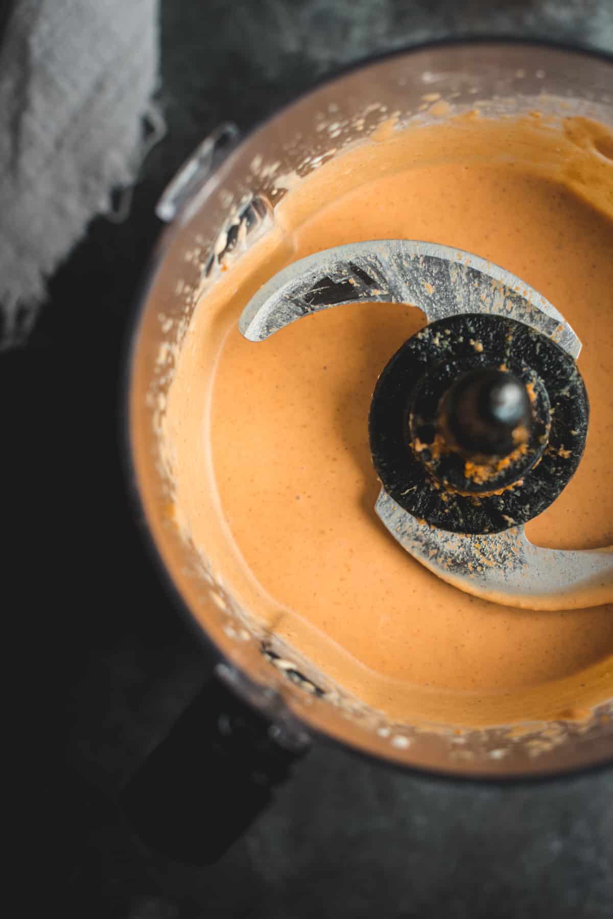 View from above of the inside of a food processor, containing heavy cream, pumpkin puree, sugar, vanilla extract and spice, it is all fully blended.